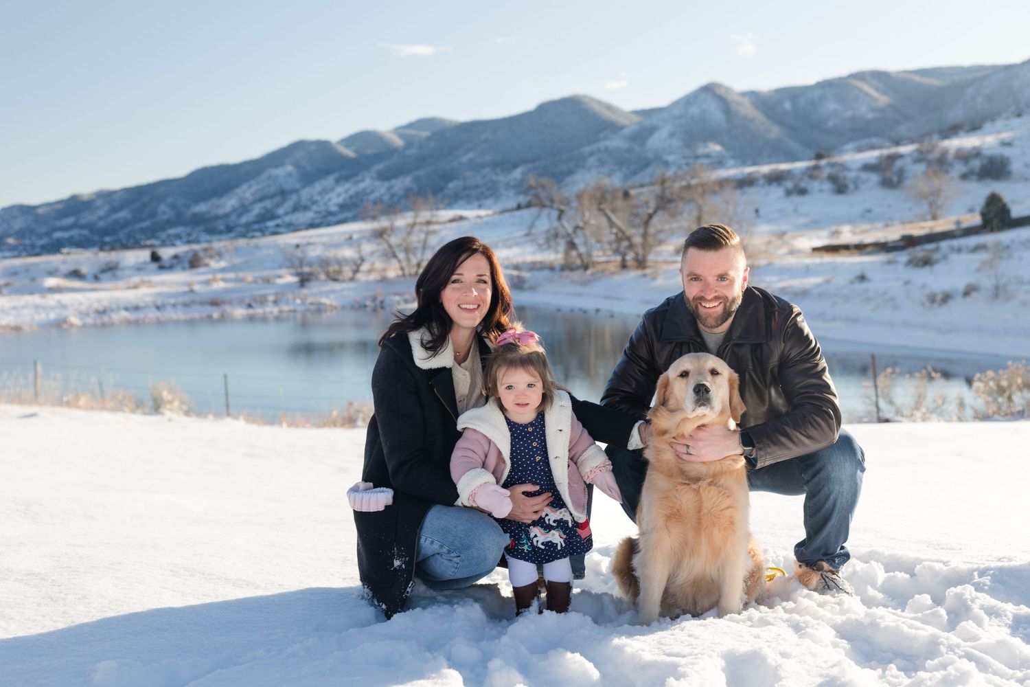 A group plays together in deep winter snow with a golden retriever dog against a scenic mountain backdrop.