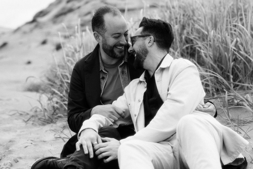 Black and white romantic beach photoshoot with couple sitting in sand dunes among tall grass.