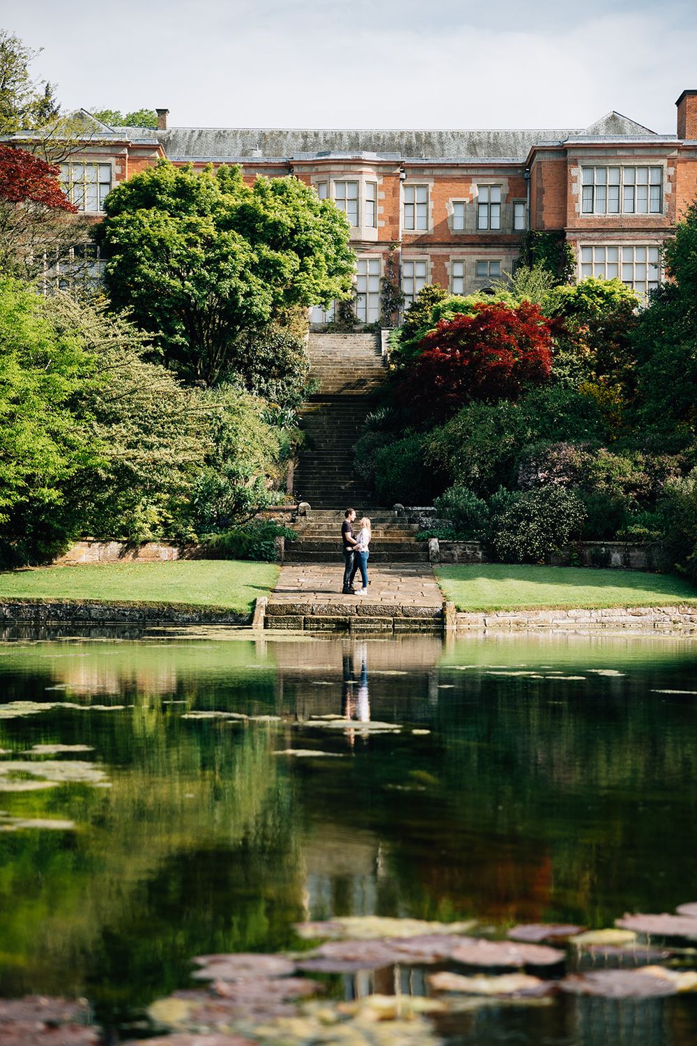A tranquil pond reflects brick buildings and lush greenery in a serene garden setting.