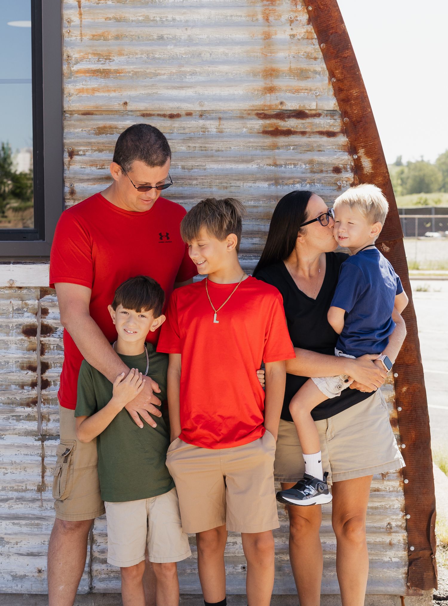 Family wearing coordinated red and khaki outfits poses against rustic metal wall outdoors.
