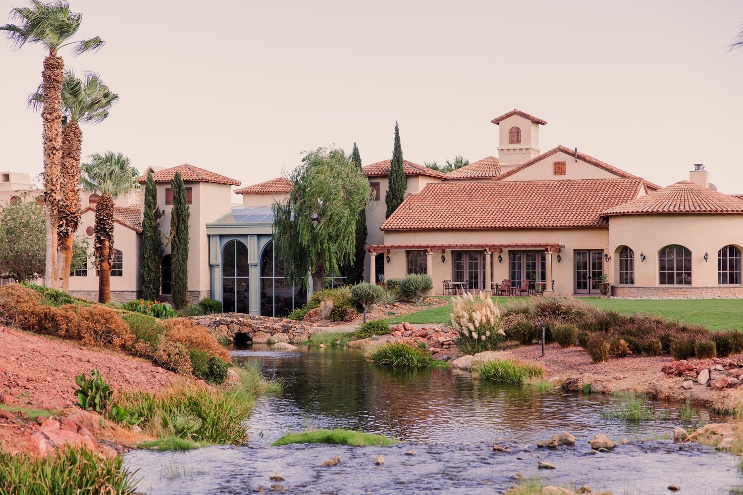 Cascata Golf Club is a tuscan-style resort with red-tile roof, lush landscaping, palm trees, and a serene pond in a desert setting.