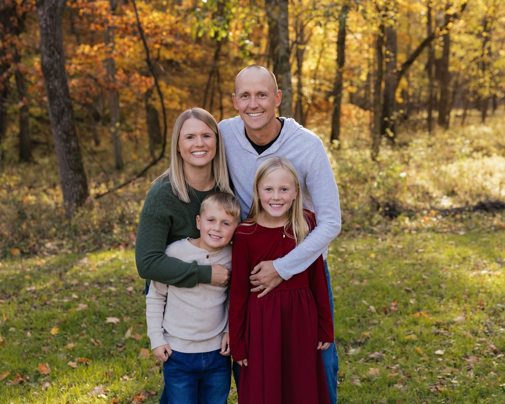 A family wearing coordinated autumn outfits poses together in a sunlit forest with golden fall foliage in the background.