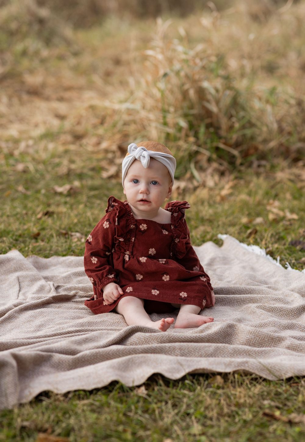 A baby in a burgundy outfit sits on a cream-colored blanket in a grassy outdoor setting.