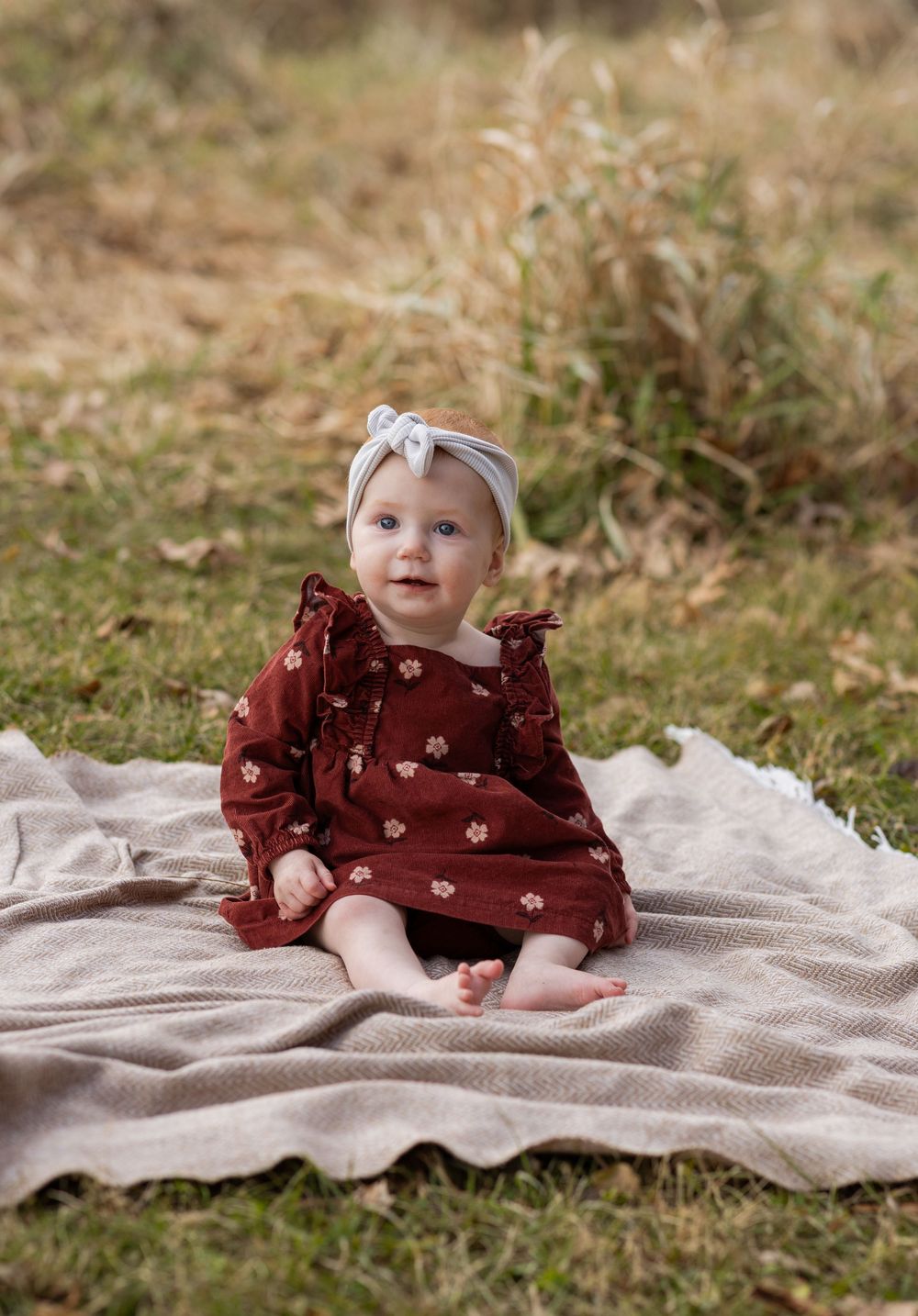 A baby in a burgundy floral dress sits on a beige blanket in a grassy outdoor setting.