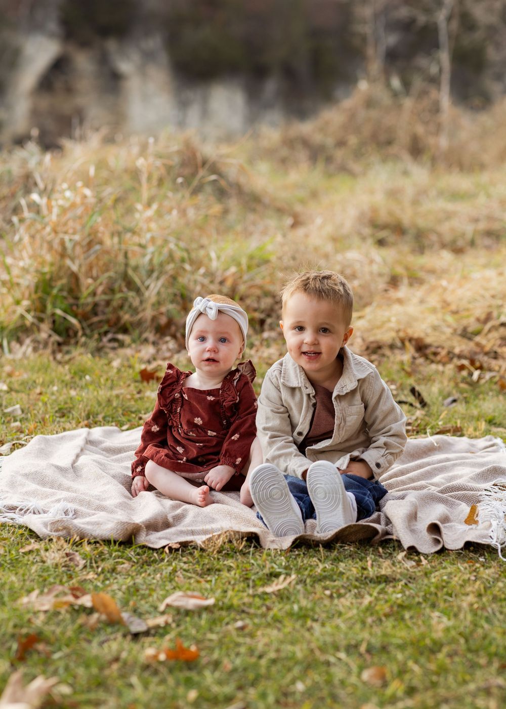 Two children laying on a blanket outdoors in autumn with fallen leaves around them.