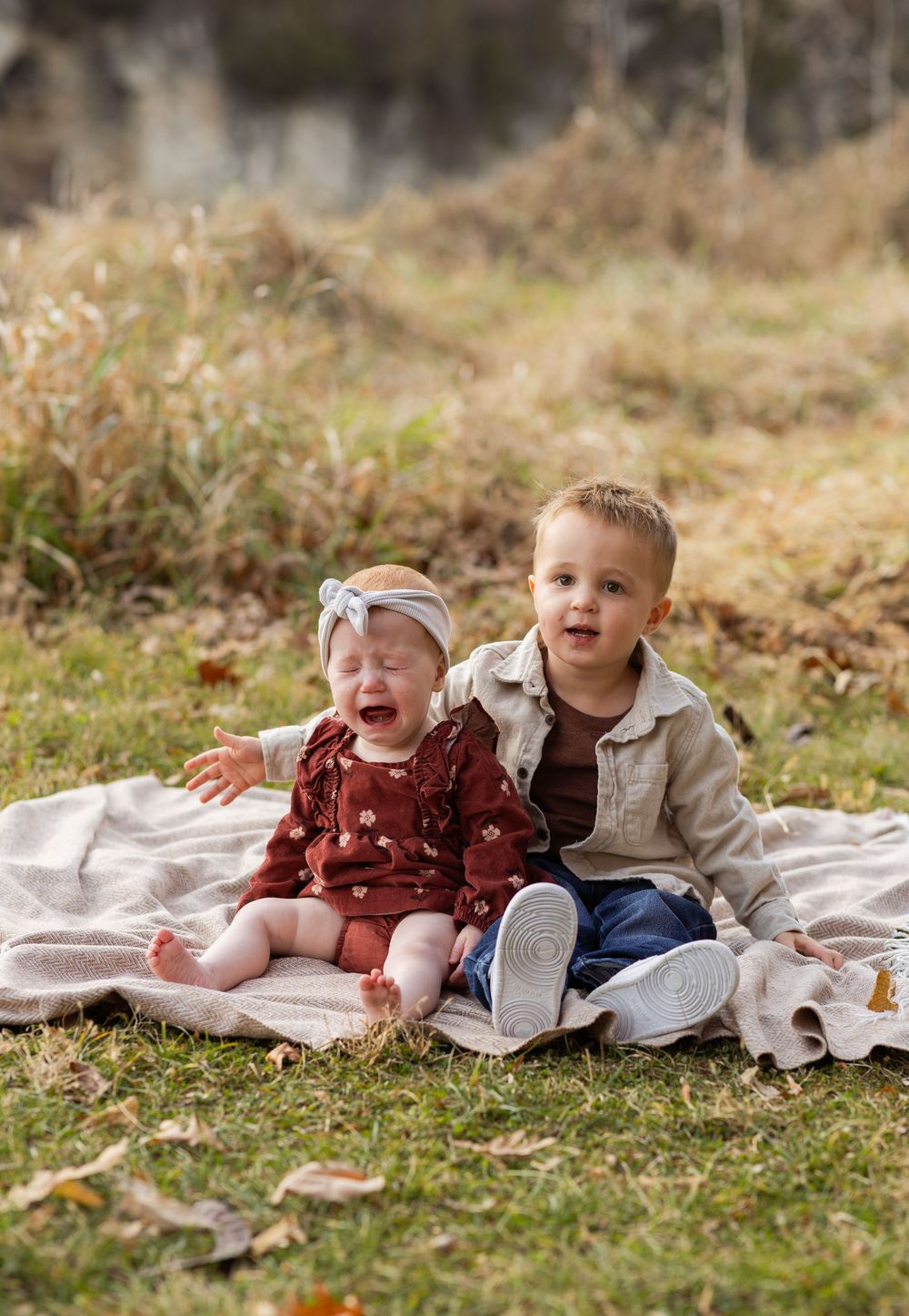 Two young children laugh and play together while lying on a white blanket outdoors in autumn.
