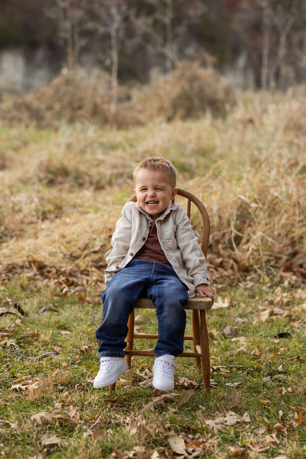A child sits on a wooden chair outdoors in autumn, wearing a beige jacket and jeans among fallen leaves.