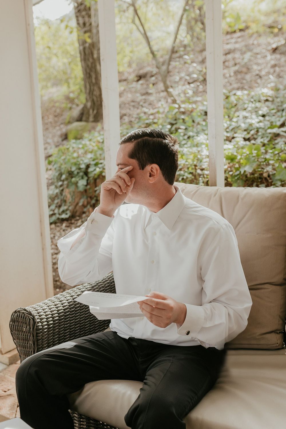 Groom in white dress shirt and dark pants wipes away tears on a wicker porch sofa with cushions at The Oaks at Duncan Lane.