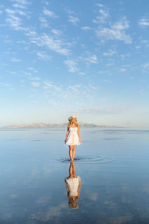 Person in white dress standing on reflective water surface with blue sky backdrop.
