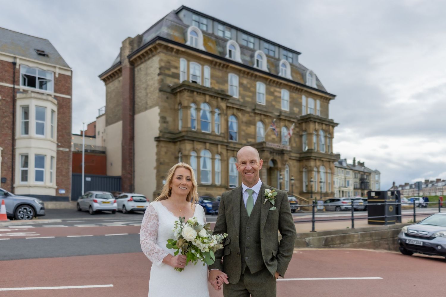 Wedding party poses outside historic brick building with cloudy sky in the background. The Grand hotel Tynemouth