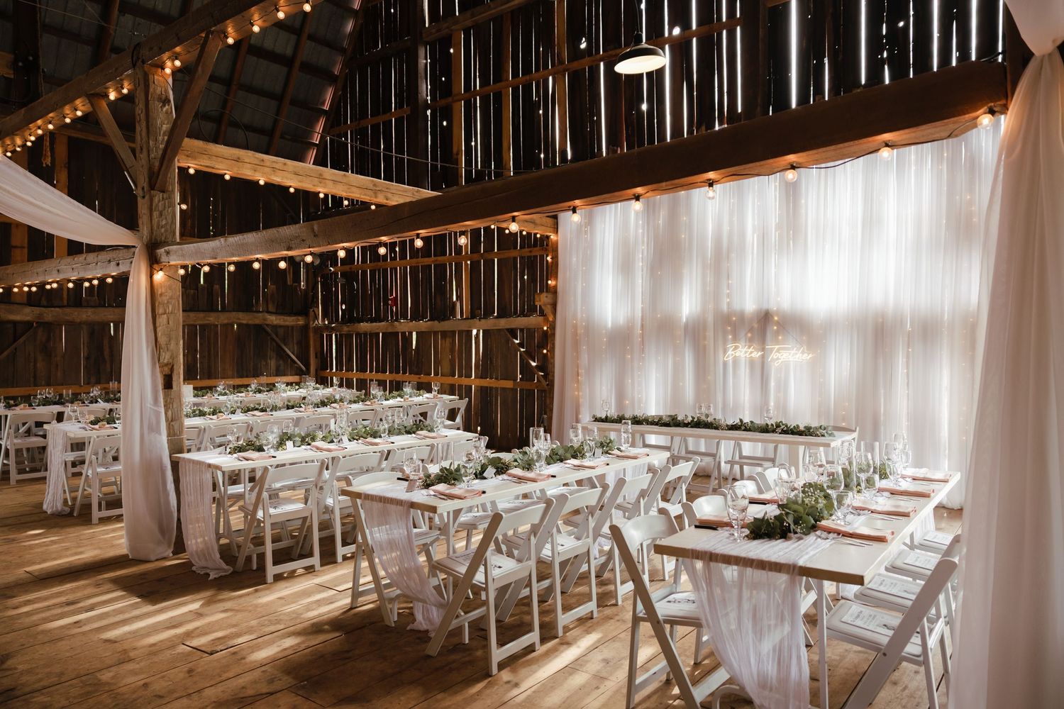 Rustic barn wedding reception setup with white tables, string lights and flowing fabric drapes creating an elegant atmosphere.