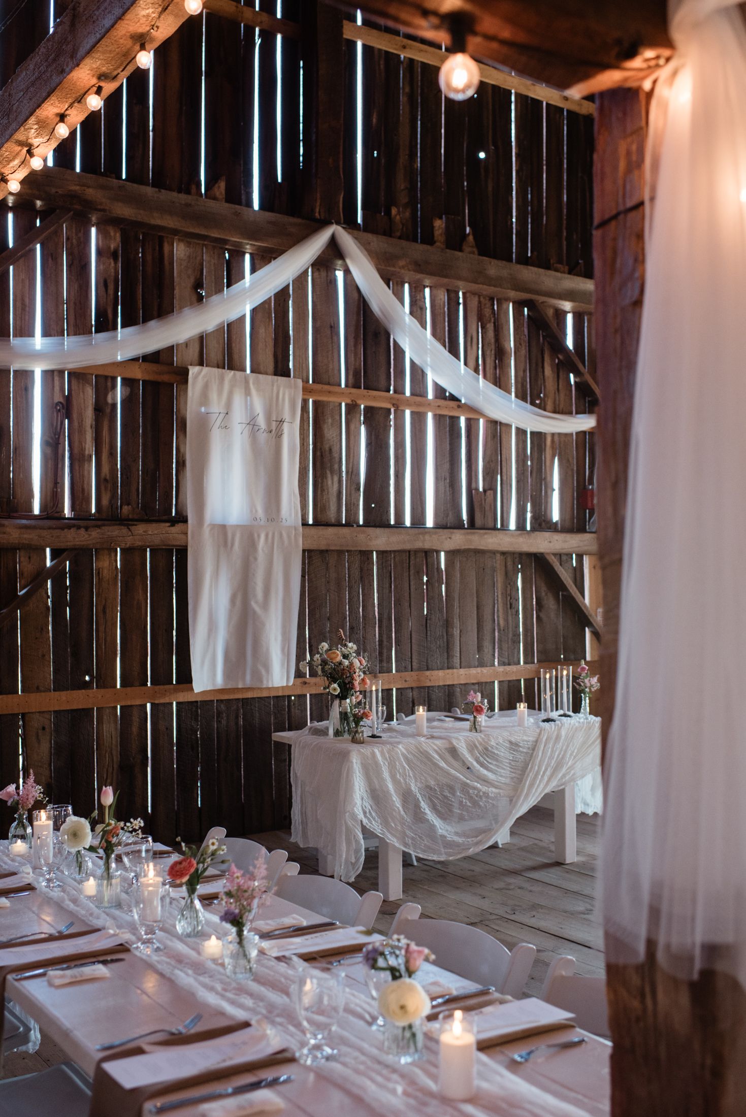 Rustic barn wedding reception setup with white drapes, wooden beams, and elegant table settings in natural light.