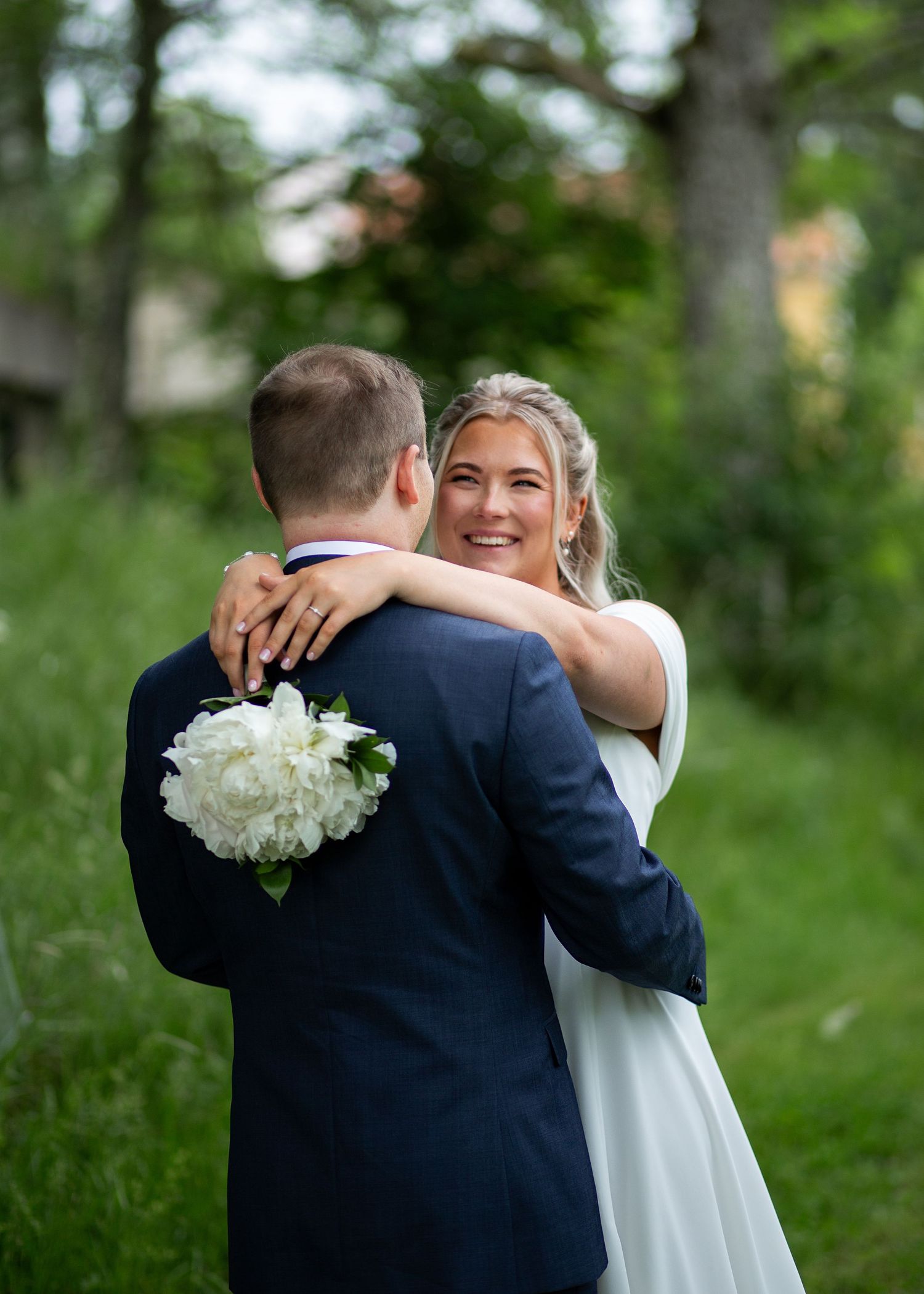Groom lifts smiling bride in white dress in a sunny green meadow with trees.
