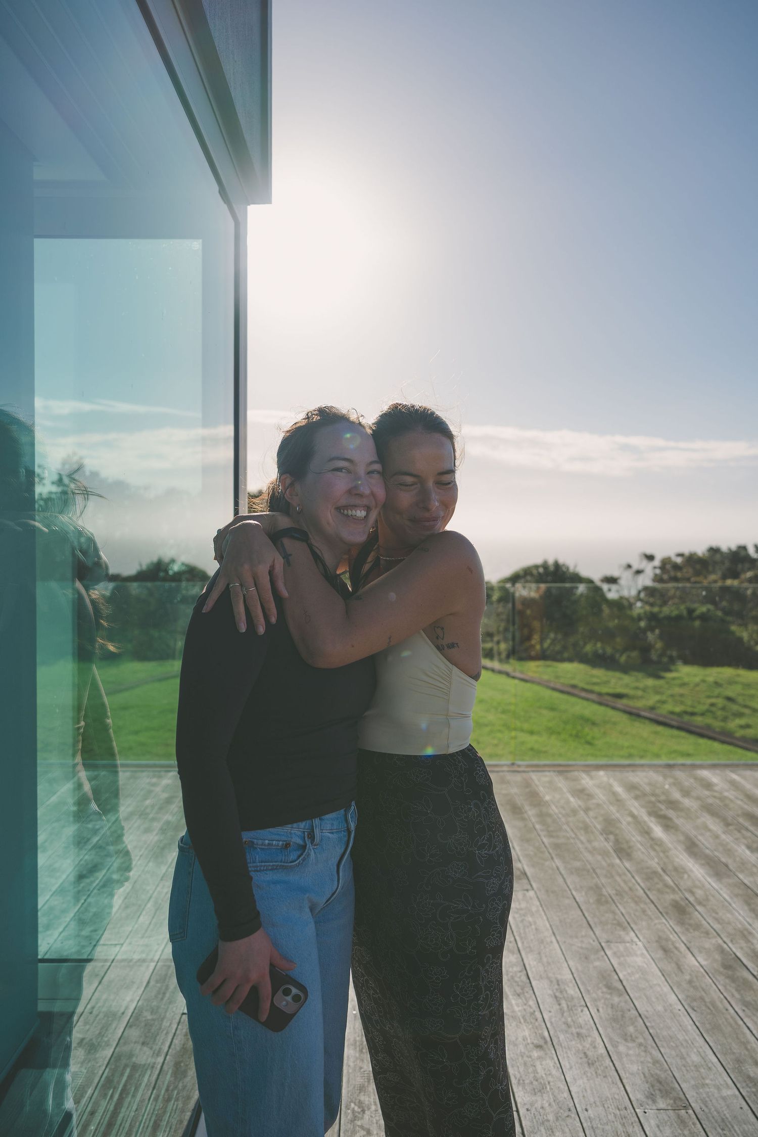 Nami embracing and laughing together on sunny deck with scenic green hillside and ocean backdrop.