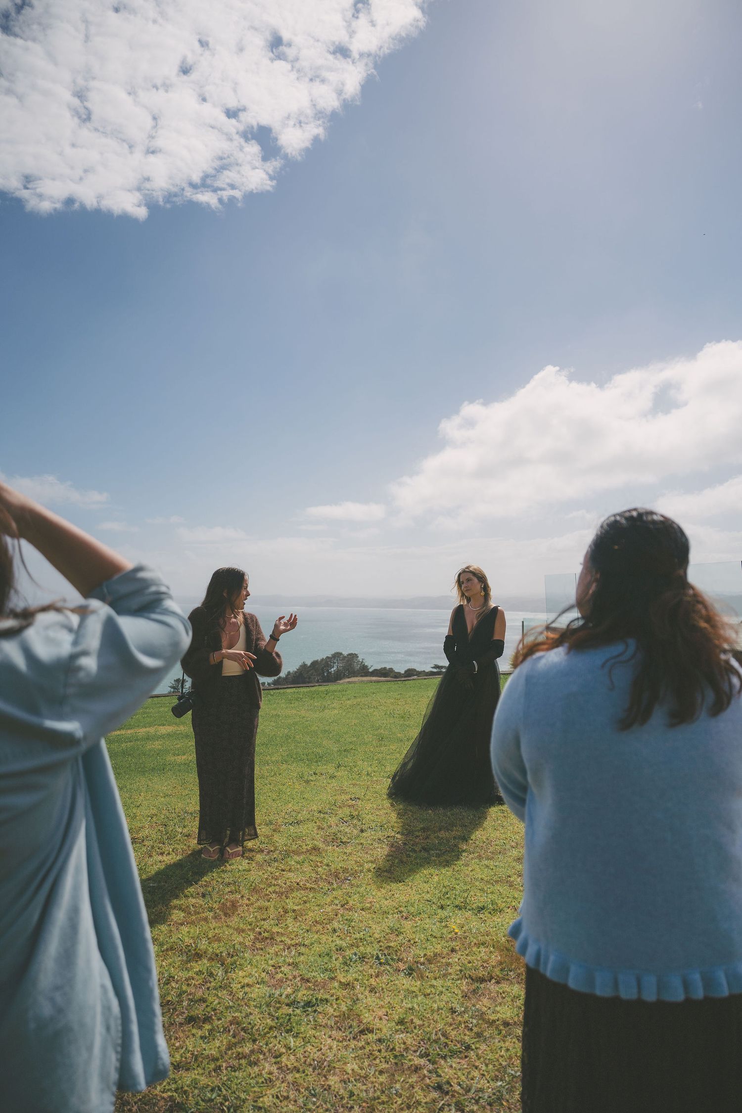 Group of women outdoors on a grassy clifftop photographing a model in a black dress by the ocean.