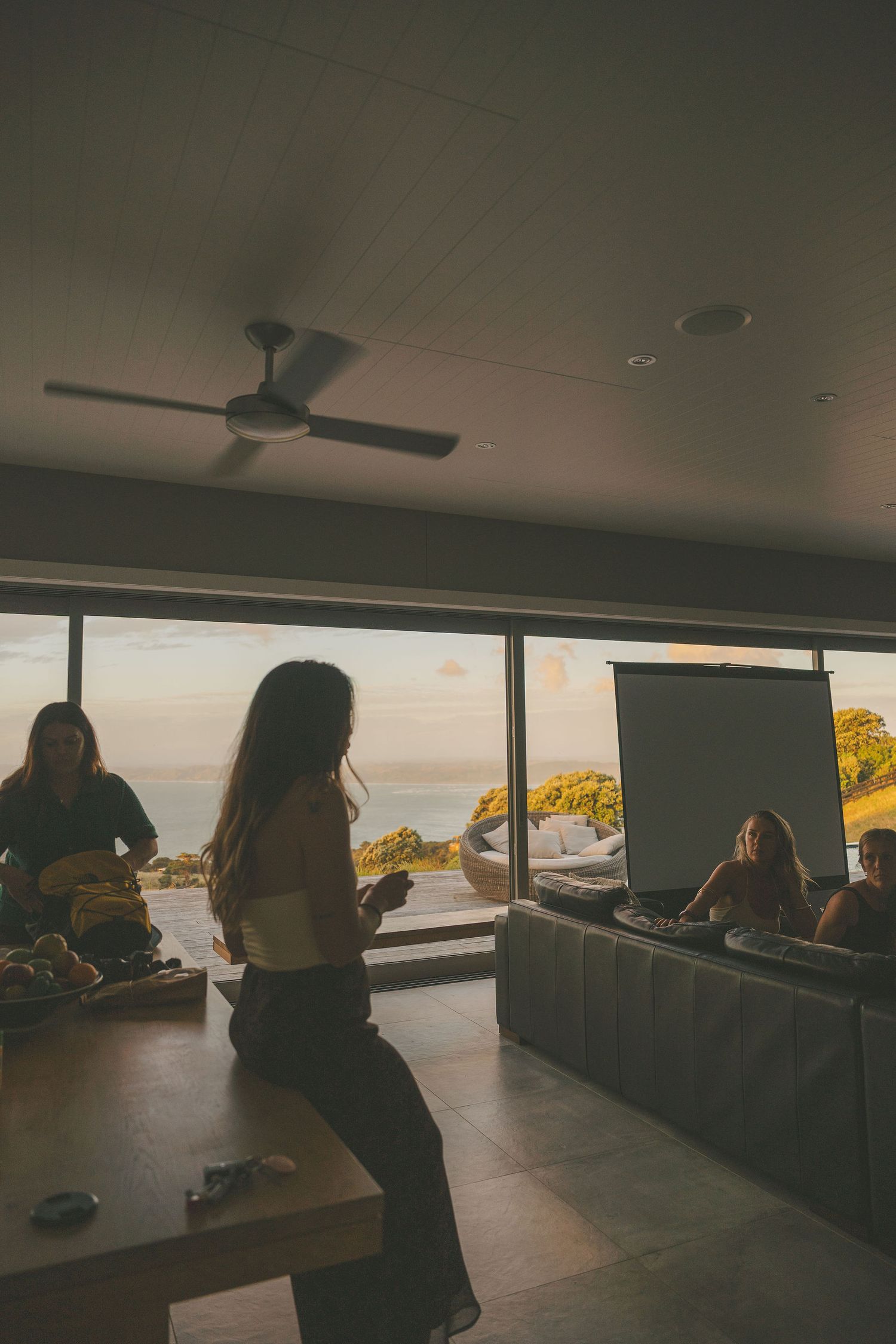 Woman perches on table during indoor sunset gathering with panoramic ocean views through large windows.