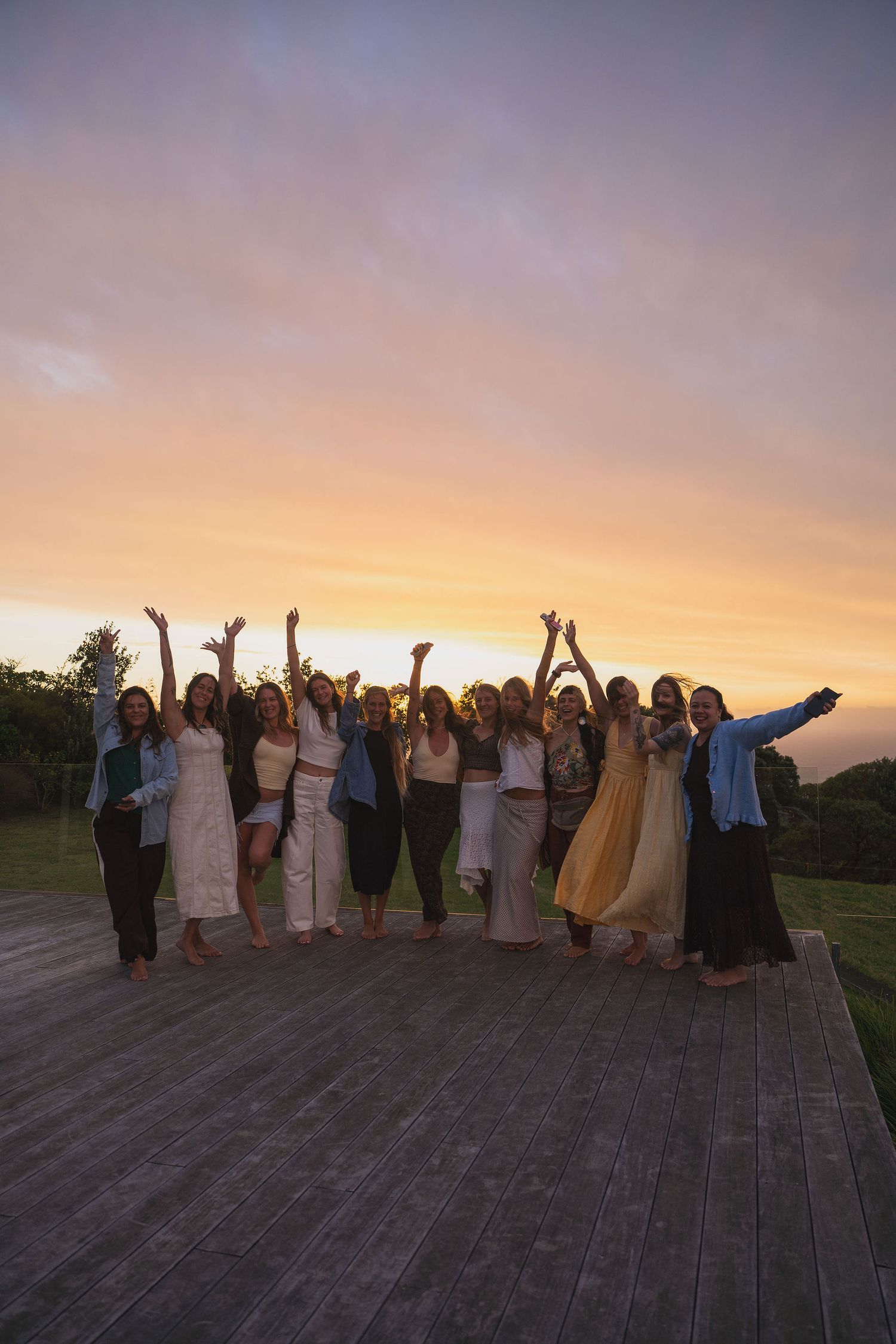 Group of women celebrating with raised arms on wooden deck during vibrant sunset with colorful sky.