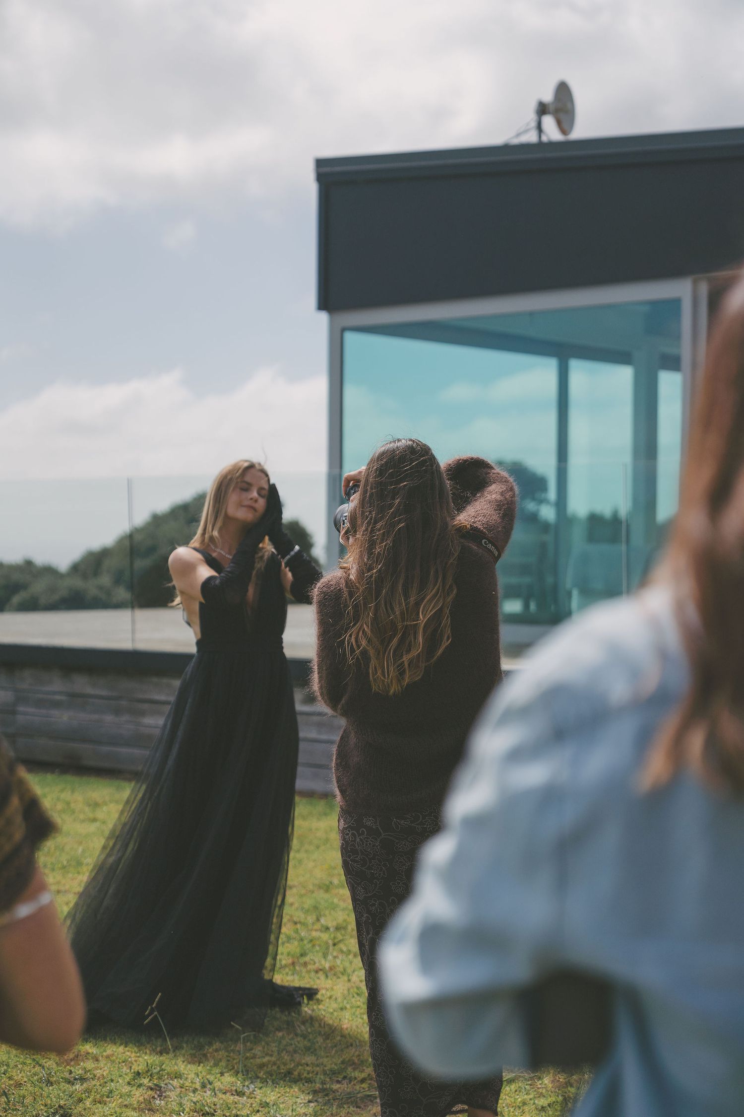 Photographer directing a model in a black gown outside a modern glass building on a cloudy day.
