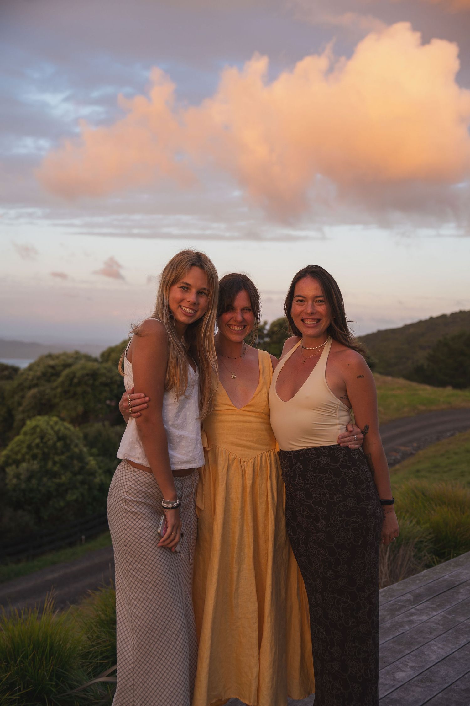 Three smiling women pose together on wooden deck during golden hour sunset with dramatic pink clouds.