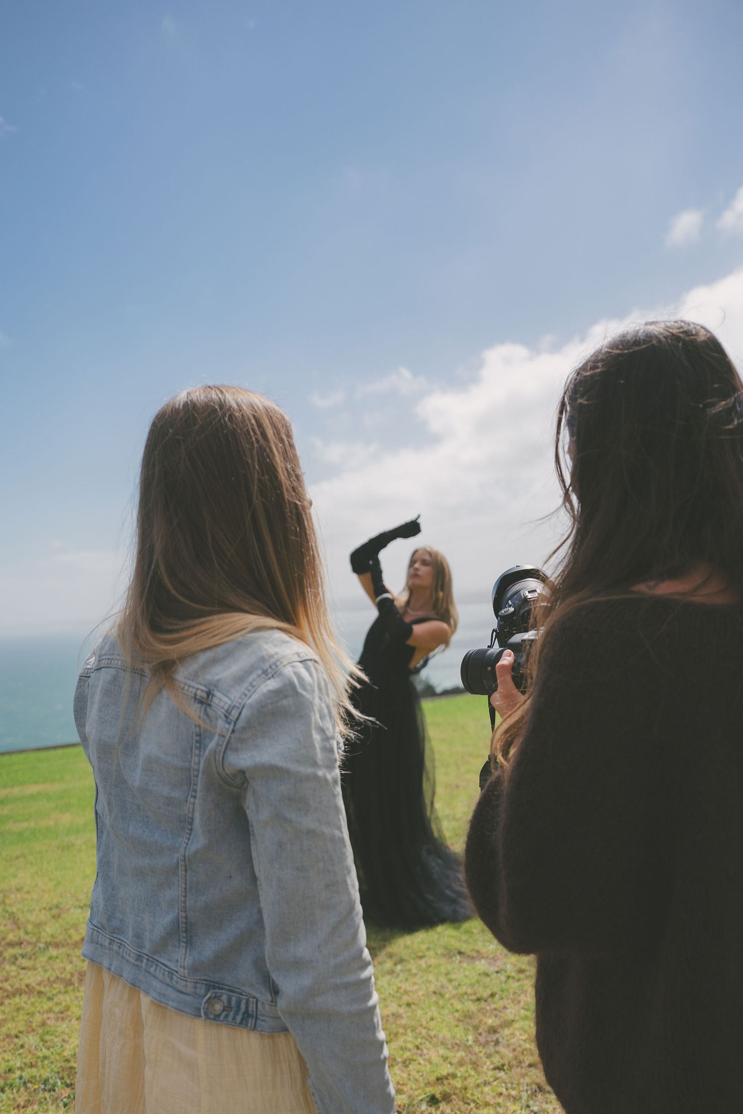 Photography students photographing a model in a flowing black dress on a scenic ocean clifftop.