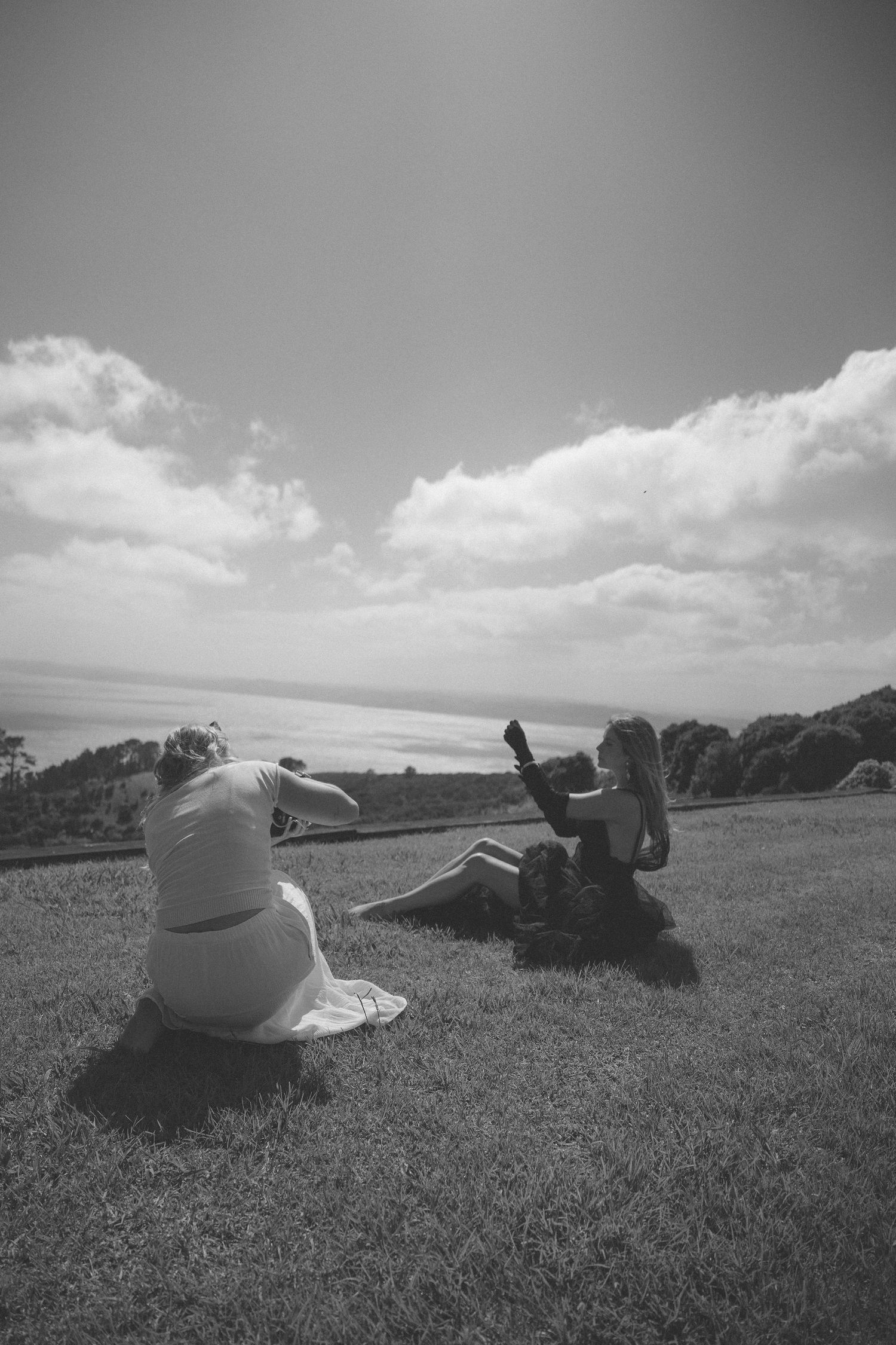 Two women sit on a grassy hillside with ocean views, one photographing the other in black and white.