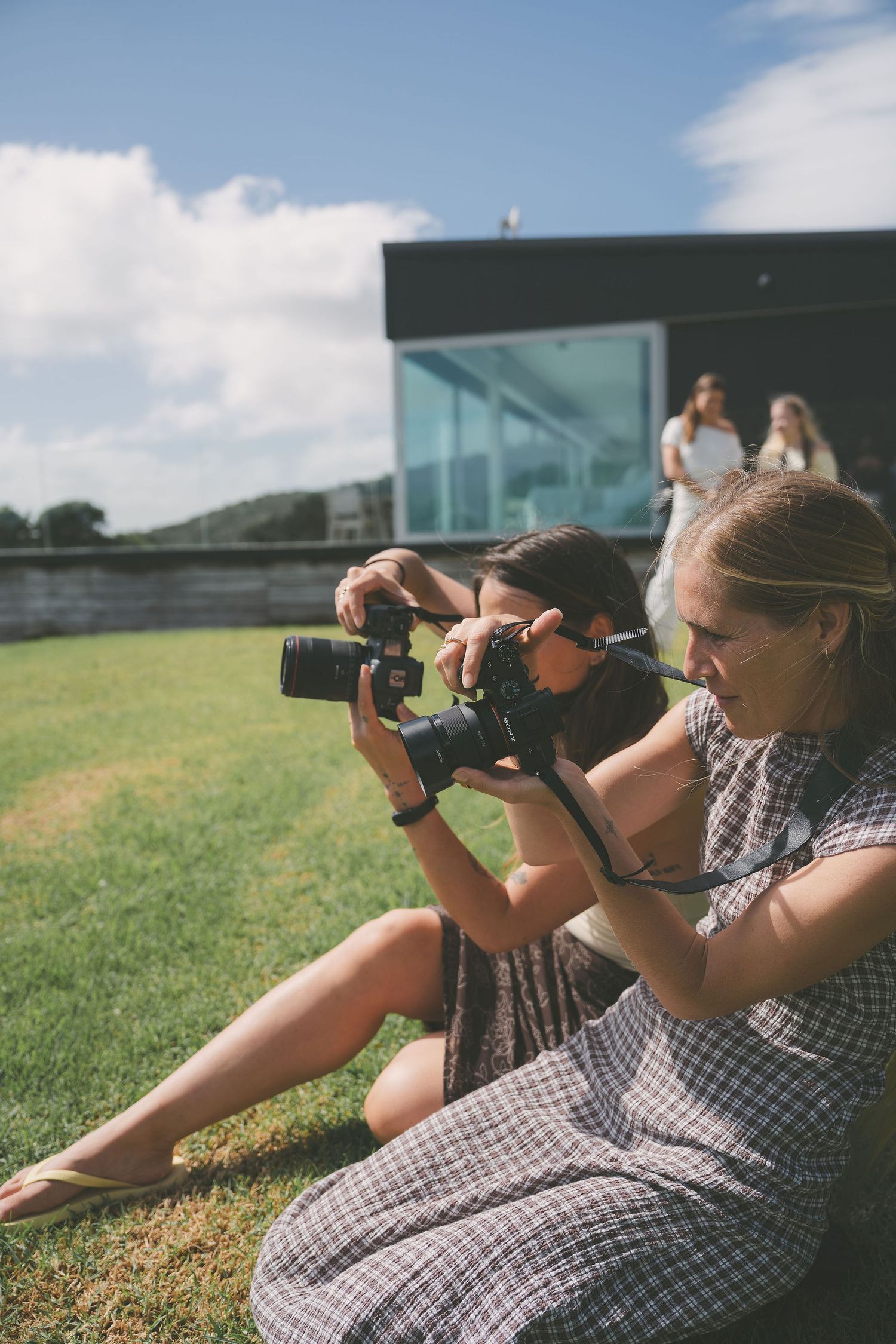 Two women sit on lawn examining DSLR camera together during outdoor photography workshop on sunny day.
