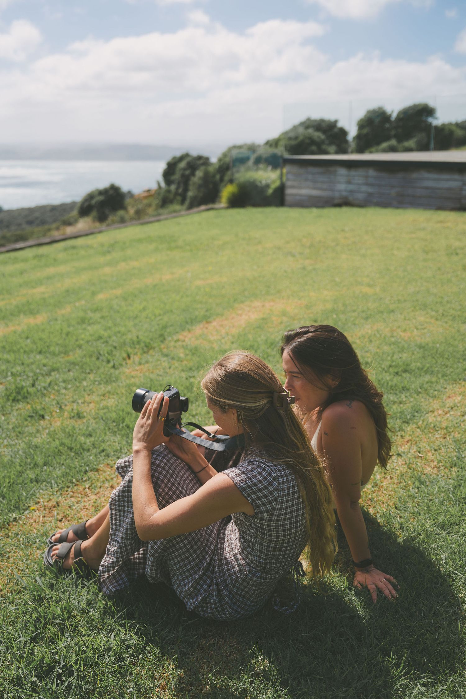 Two women sit on green grass reviewing photos on a vintage film camera with ocean views.