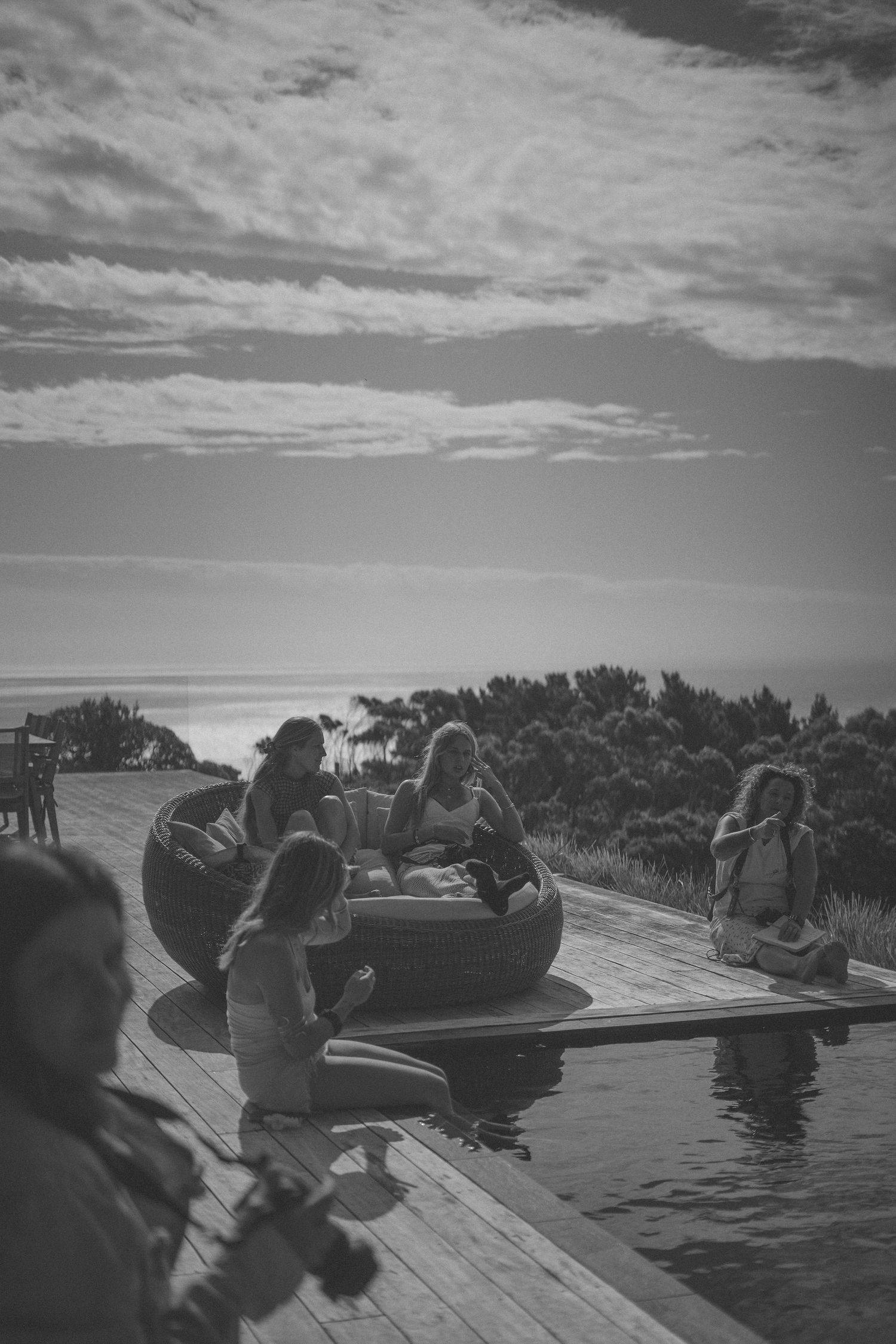 Group of women relaxing by infinity pool on wooden deck with ocean views in black and white.