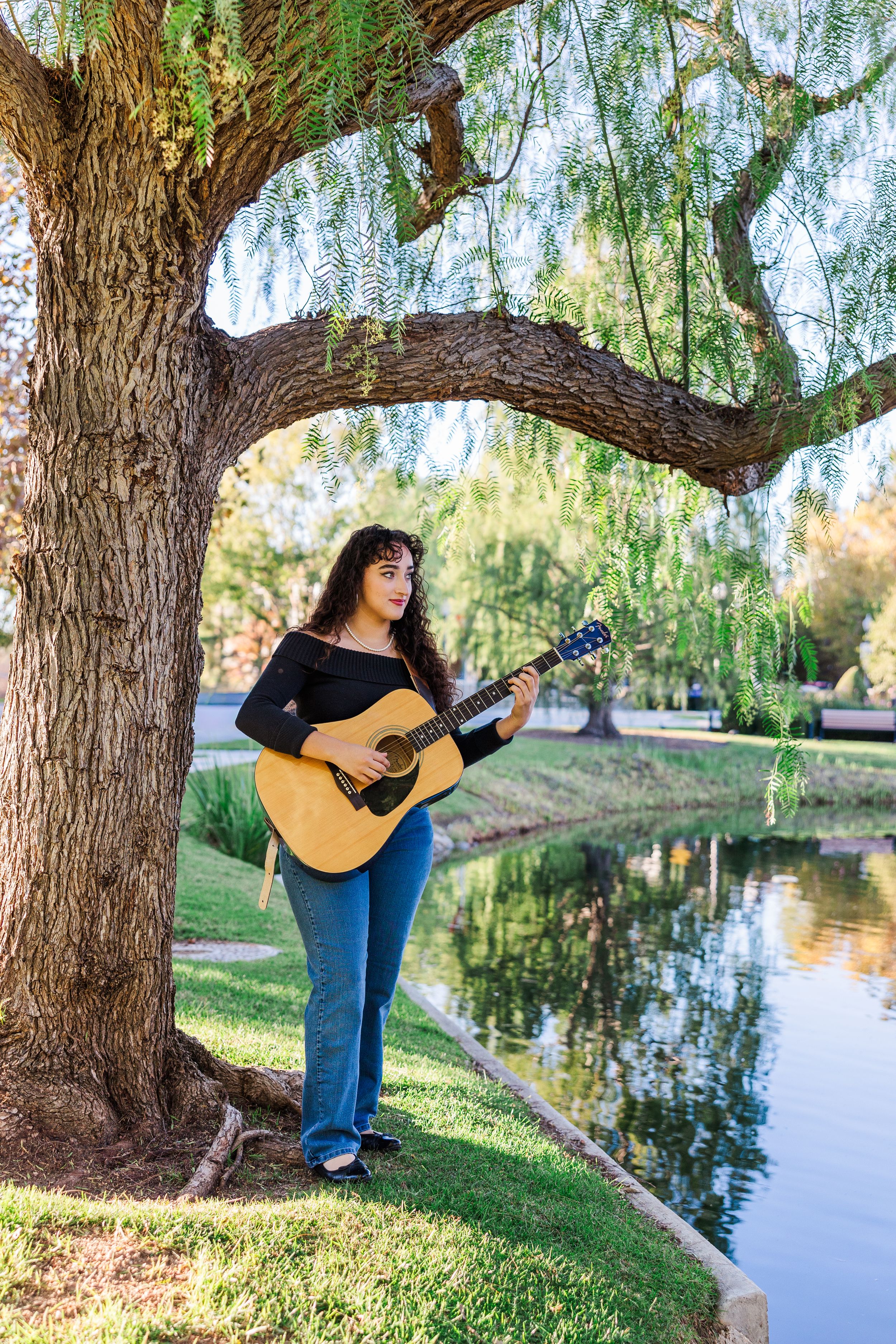 A musician plays acoustic guitar under a weeping willow tree beside a tranquil pond on a sunny afternoon.