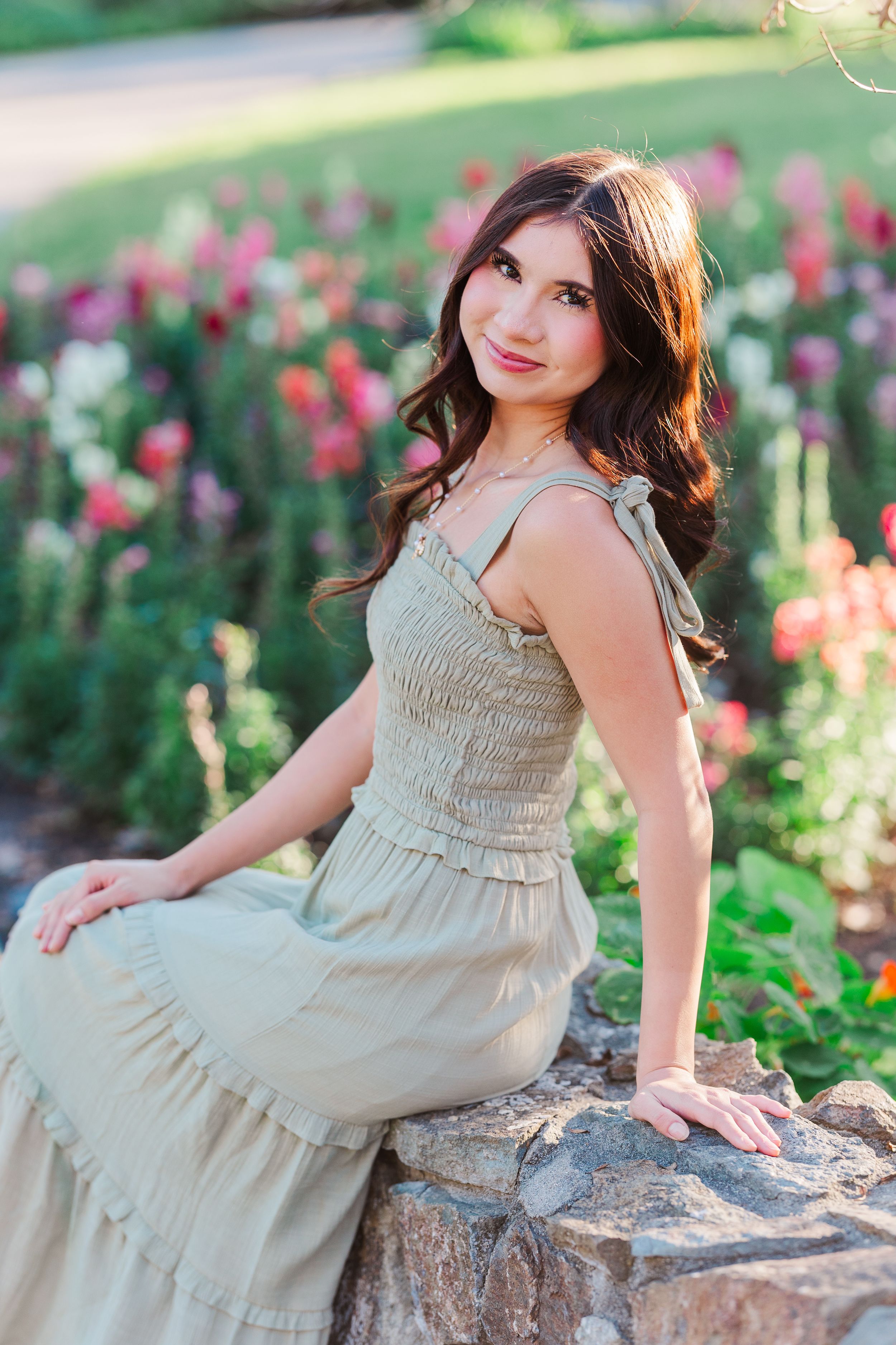 A woman in a beige smocked dress poses gracefully on a rock in a garden with blooming pink flowers.