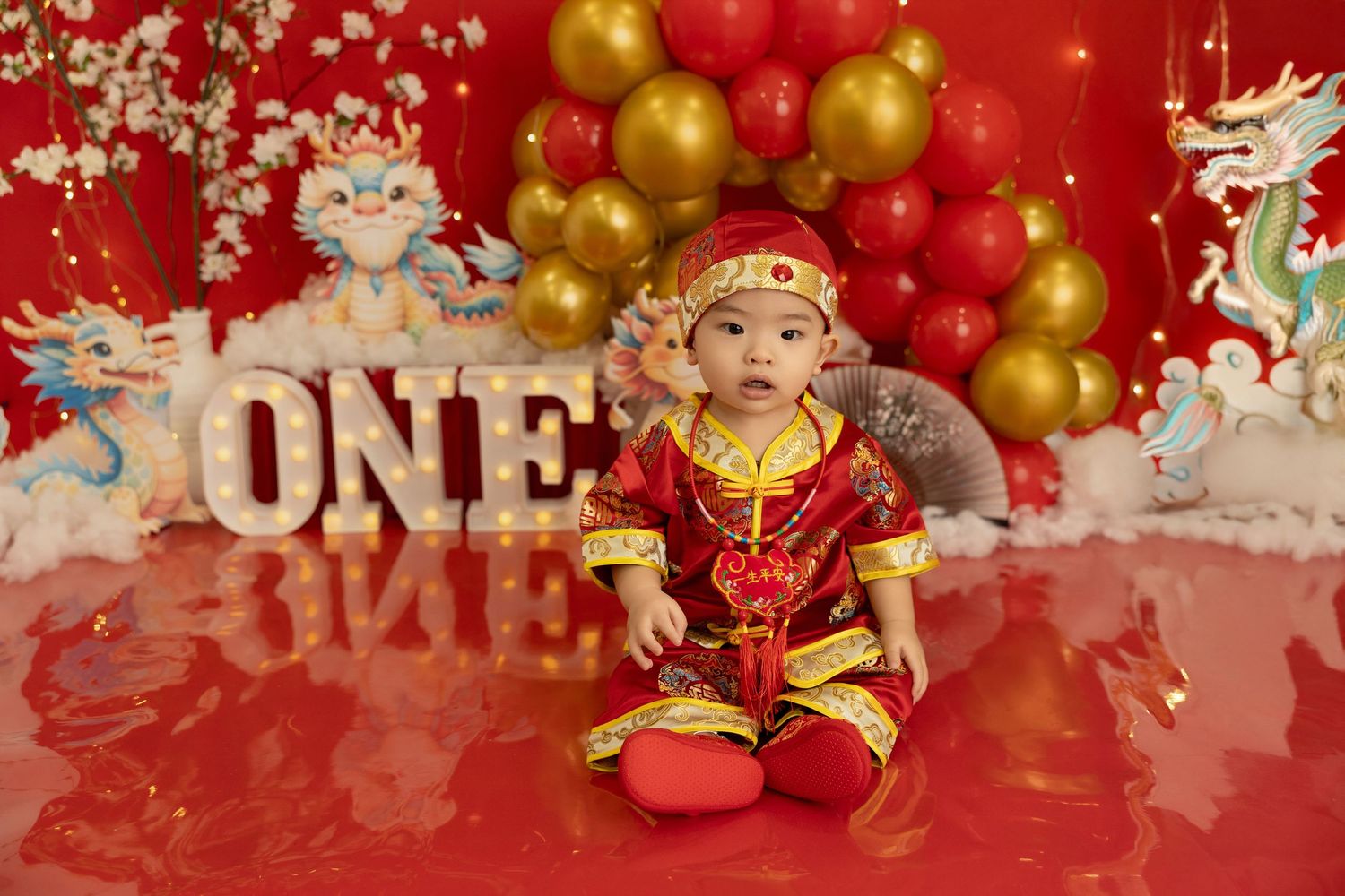 First birthday setup with traditional Chinese outfit, gold and red balloons, and decorative letters spelling ONE.
