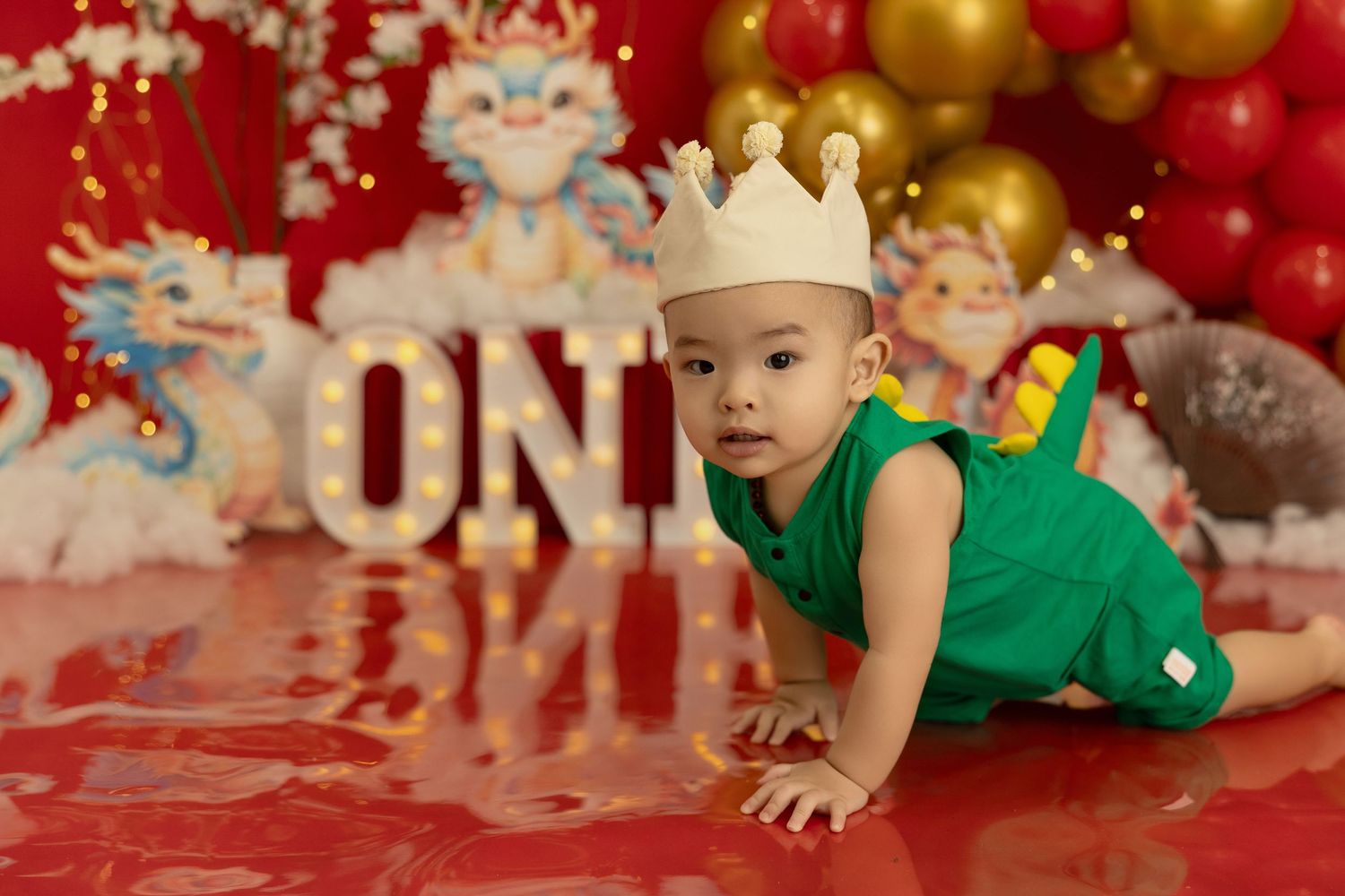 Baby in green shirt crawls on red and gold holiday backdrop during festive Christmas photo session.