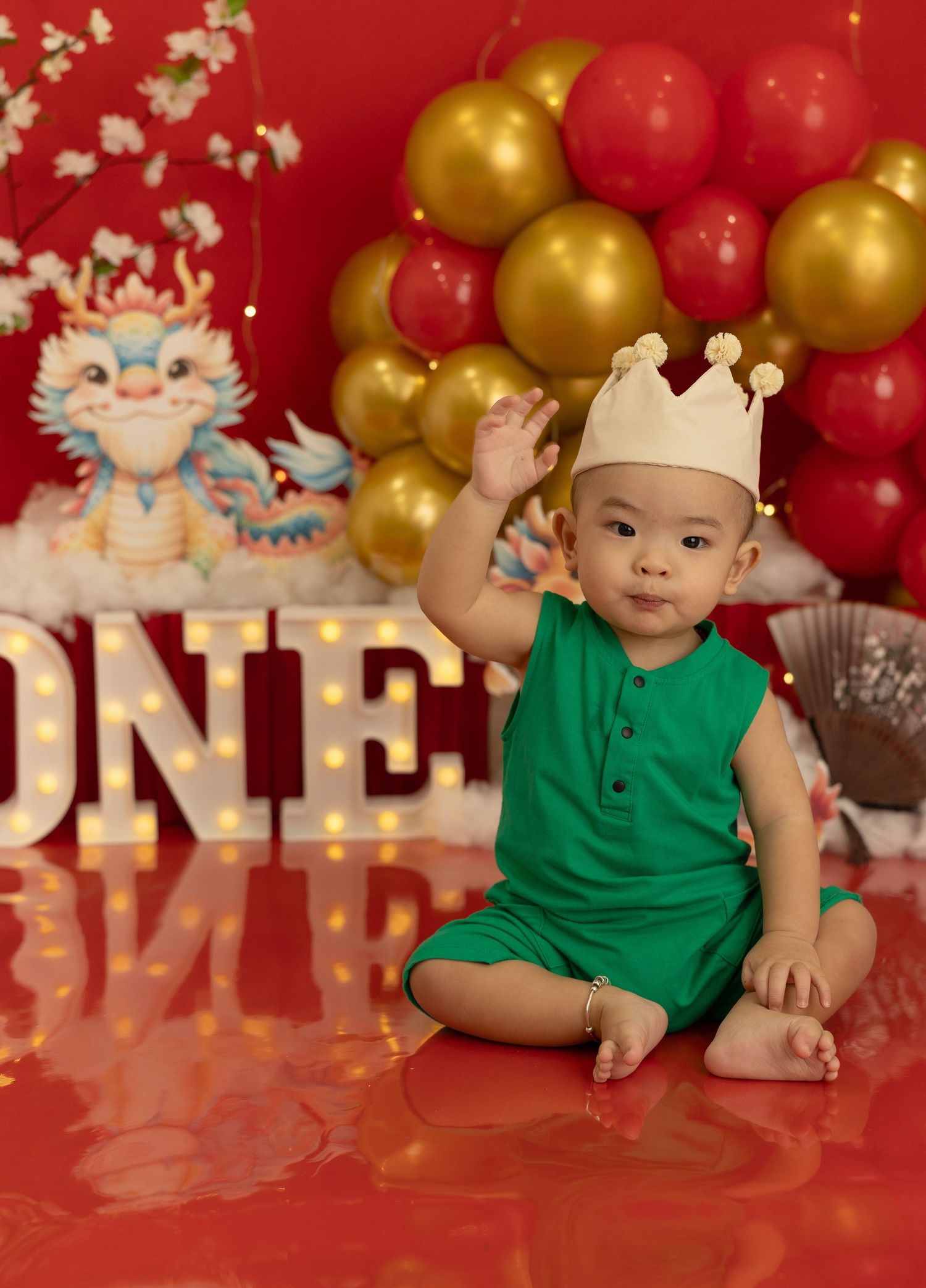 A baby in a green polo shirt poses against a festive red backdrop with gold and red balloons.