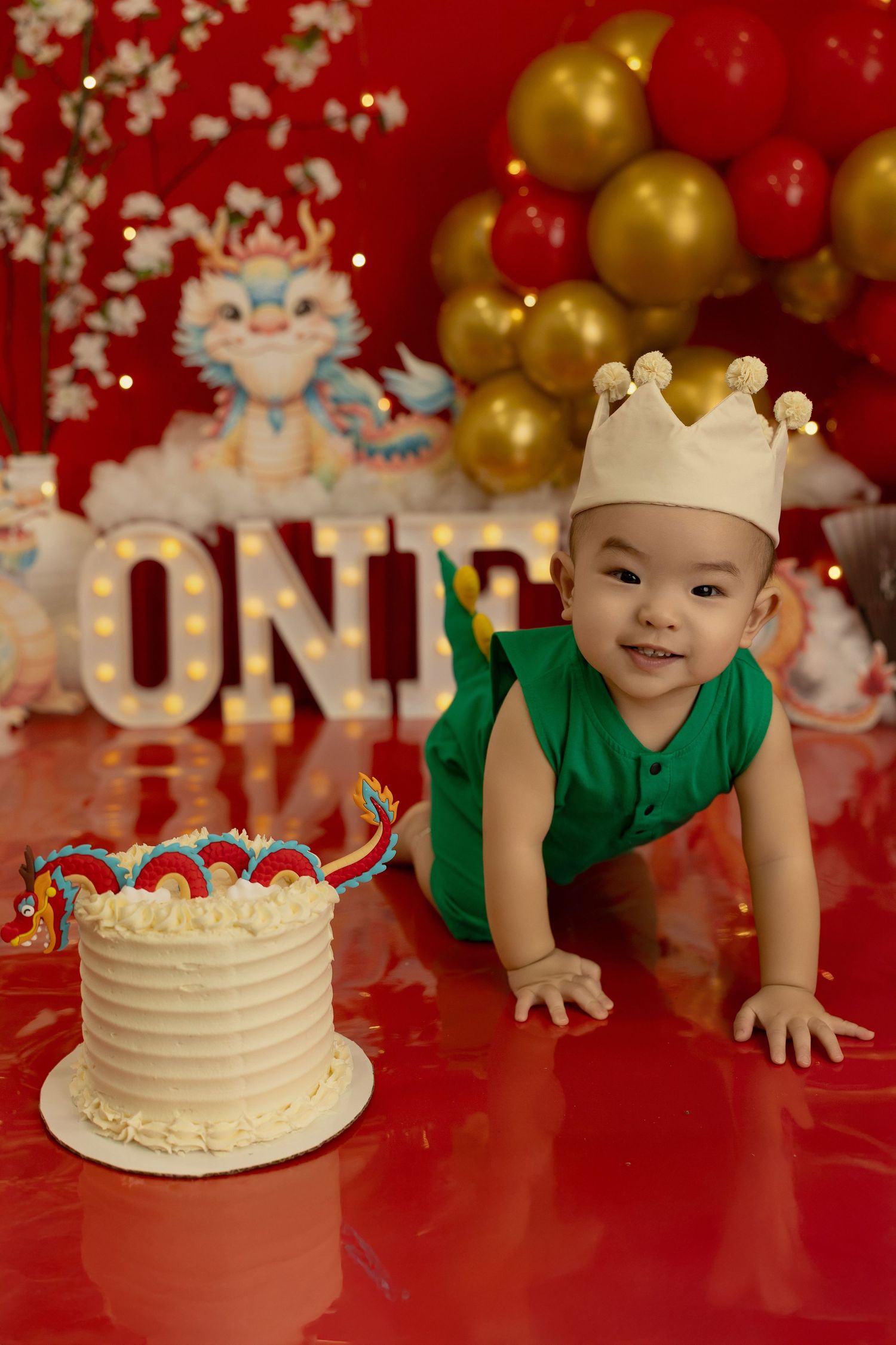 A first birthday celebration setup with a white cake and decorative balloons against a red backdrop.