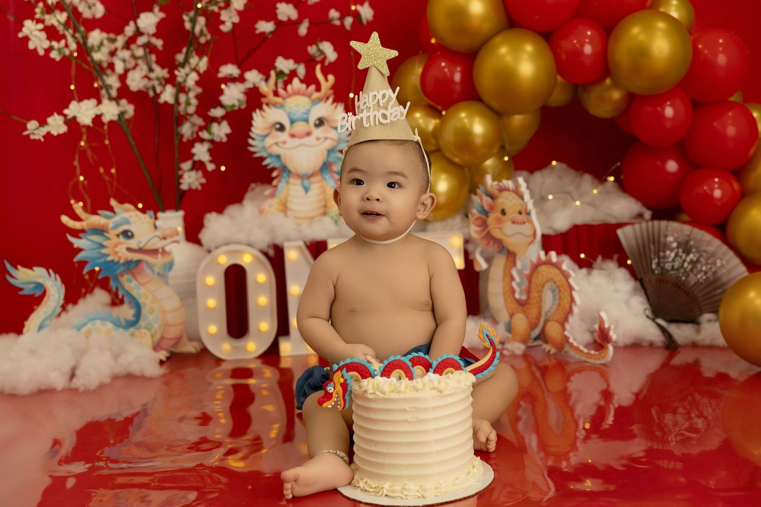 Birthday celebration setup with white cake, gold and red balloons, and festive decorations on red background.