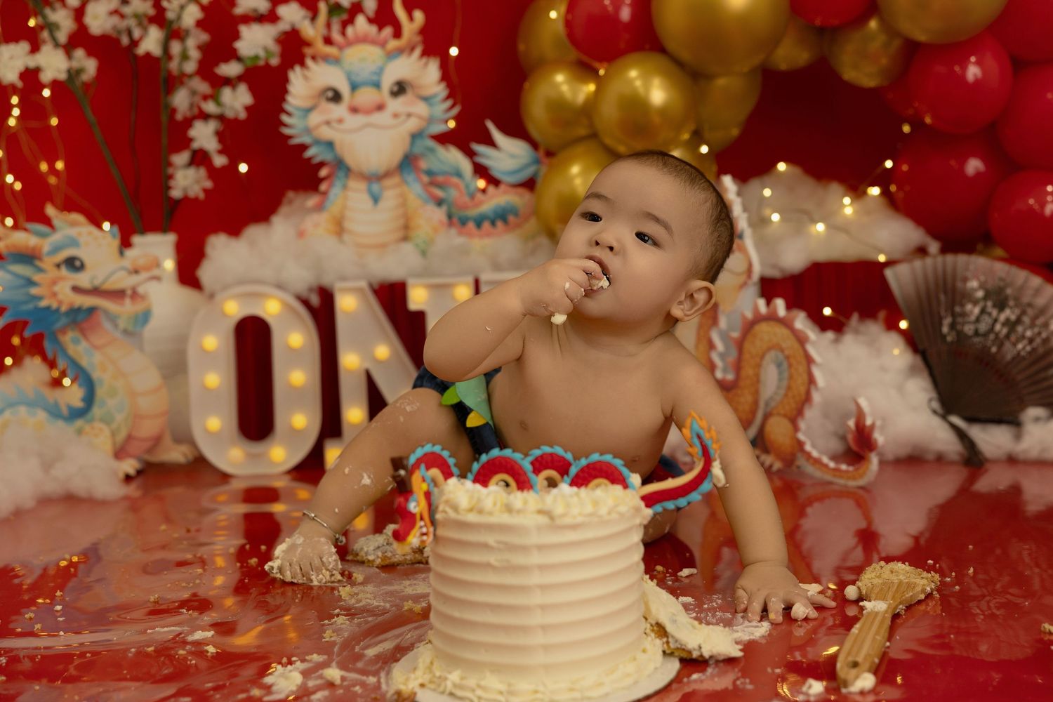 Festive cake smash setup with red and gold balloons against a red backdrop with twinkling lights.