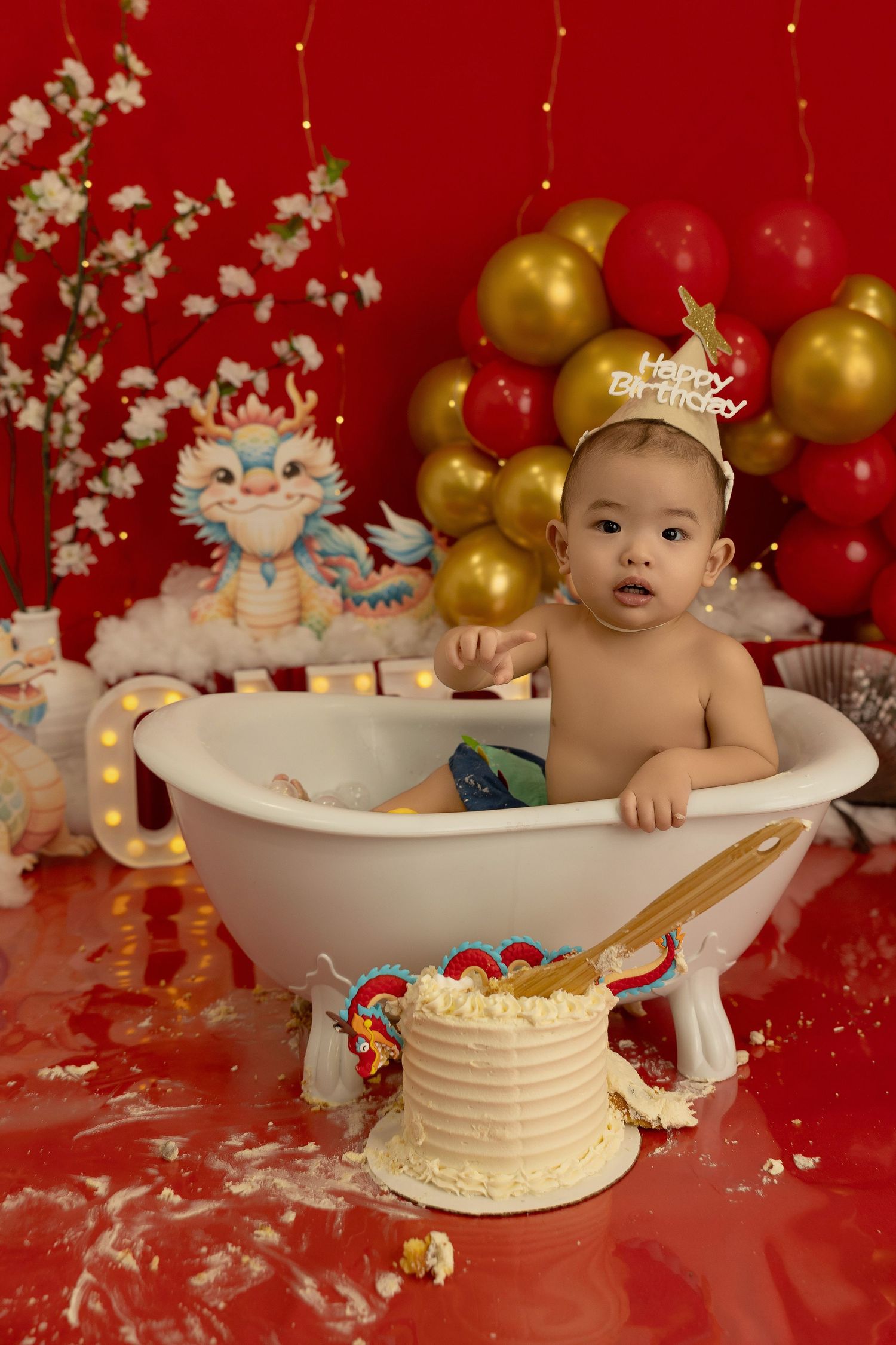 Festive first birthday scene with bathtub, birthday cake, gold and red balloons against red backdrop.