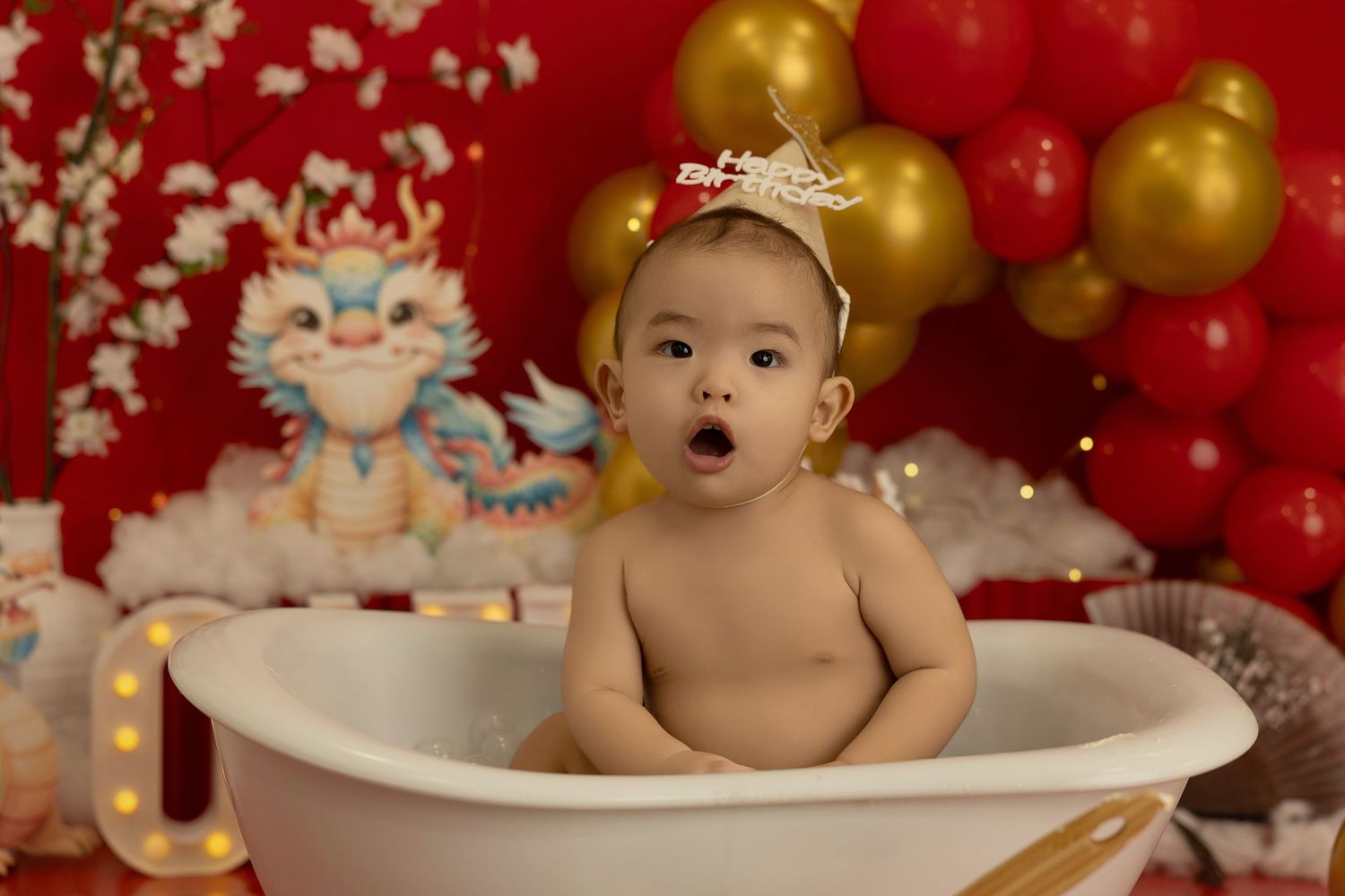 A festive bath photography setup with red and gold balloons and floral decorations in the background.