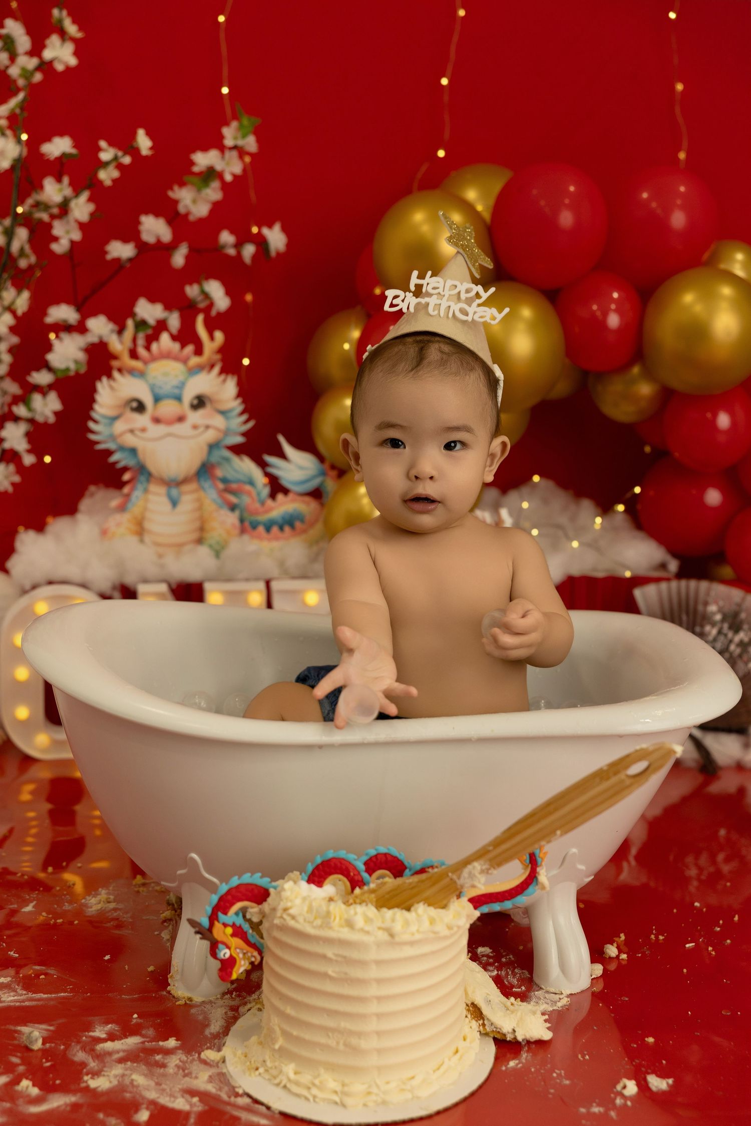 Cake smash photo session featuring a white bathtub prop and festive red and gold balloon decorations.