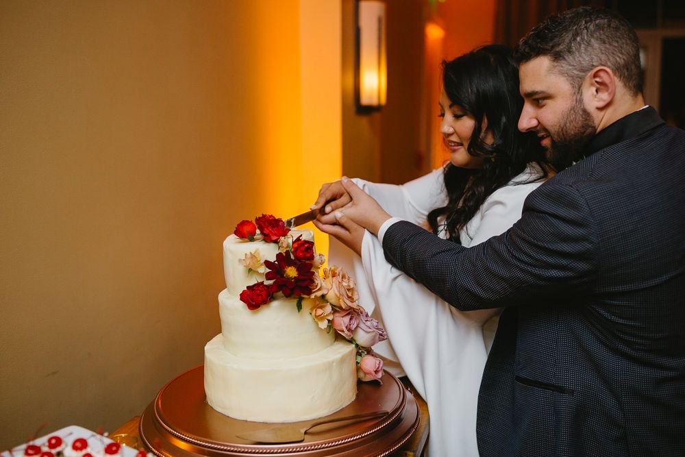 A couple cuts into their elegant white wedding cake decorated with red roses at a reception.