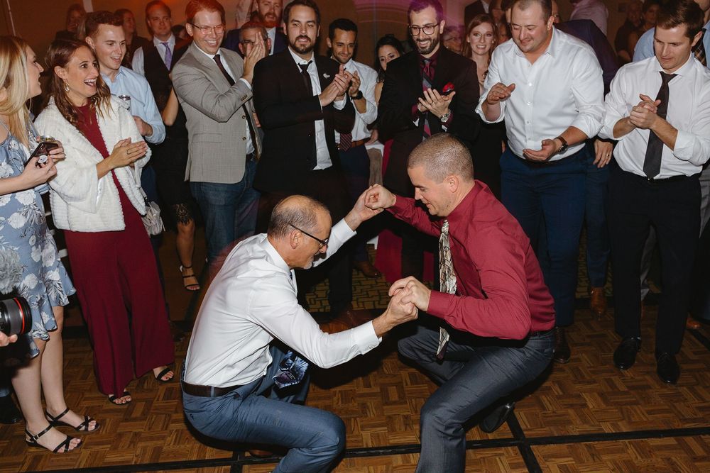 Two men dance energetically on a wooden floor surrounded by cheering wedding guests in formal attire.