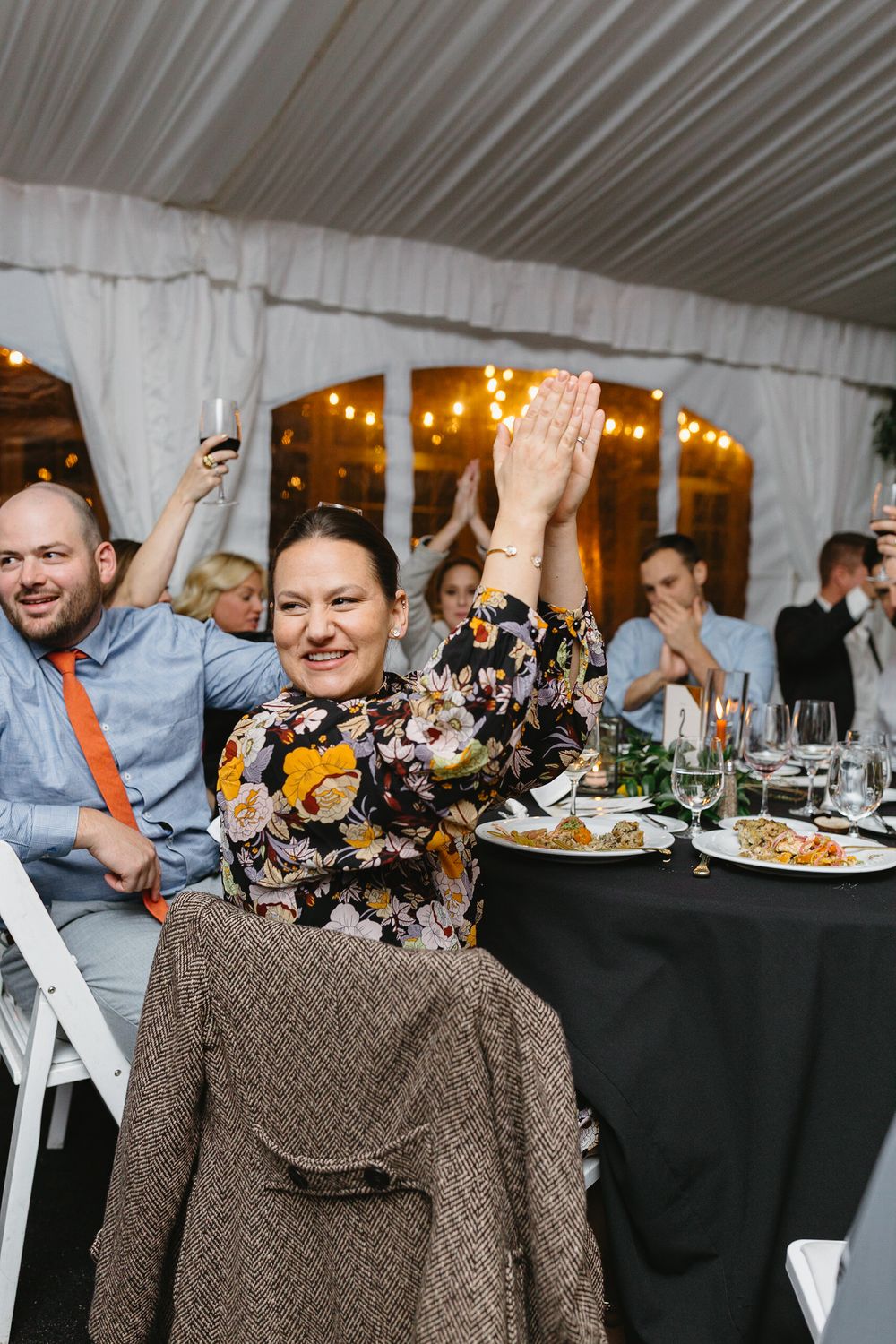 Wedding reception guests seated at tables with black tablecloths enjoying dinner and raising hands in celebration.