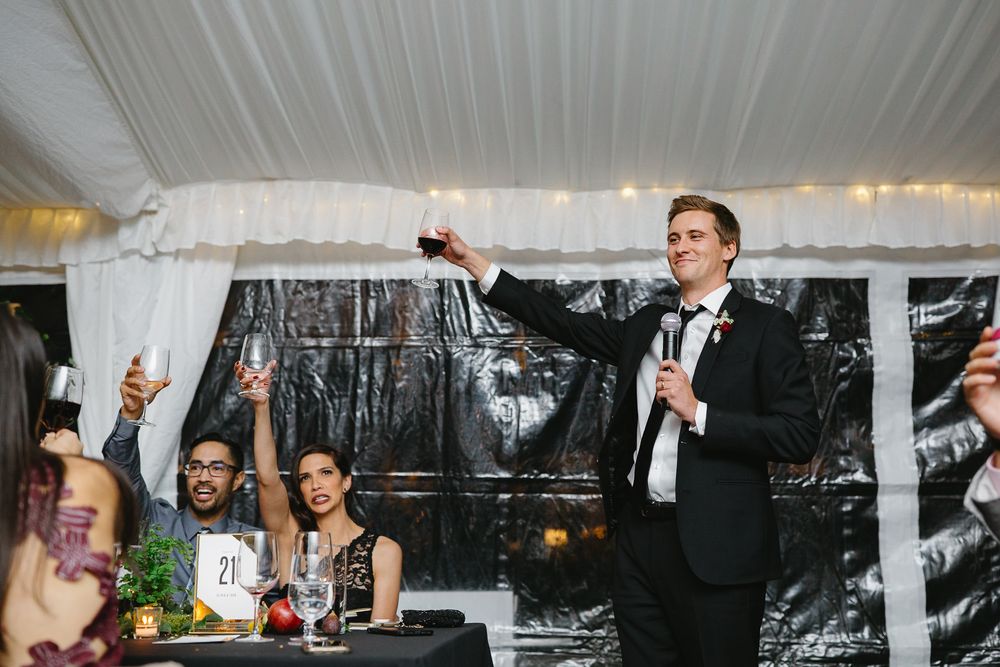 A person in a black suit gives a toast while holding a microphone at a formal event under a white tent.