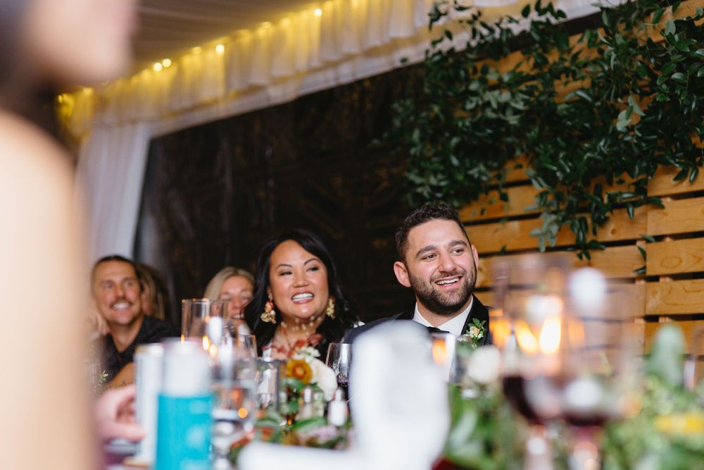 Wedding guests laugh and smile during reception dinner at rustic venue with wooden walls and greenery decor.