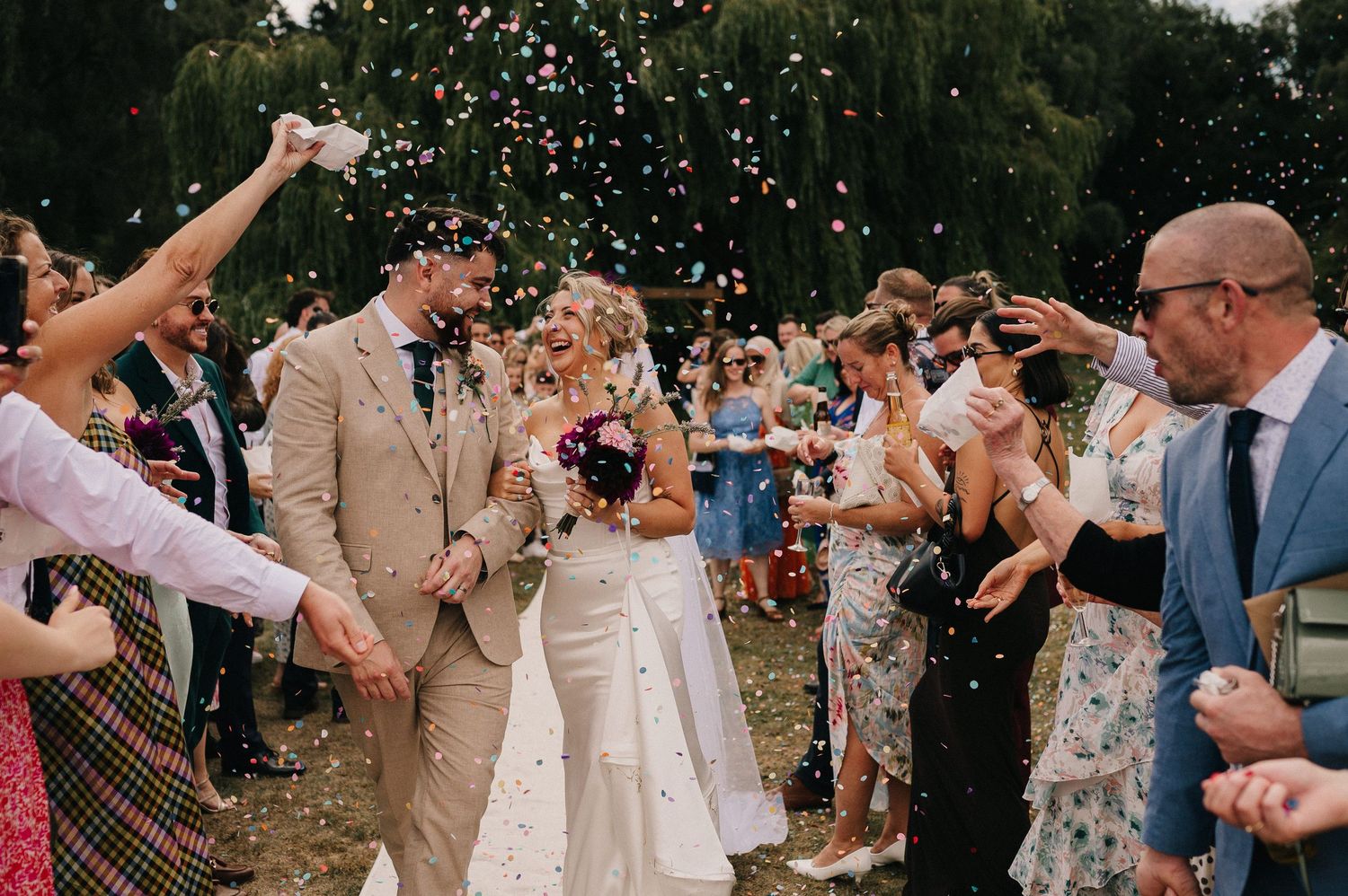 Wedding guests toss confetti over newlyweds during outdoor celebration with joyful crowd in summer garden setting.