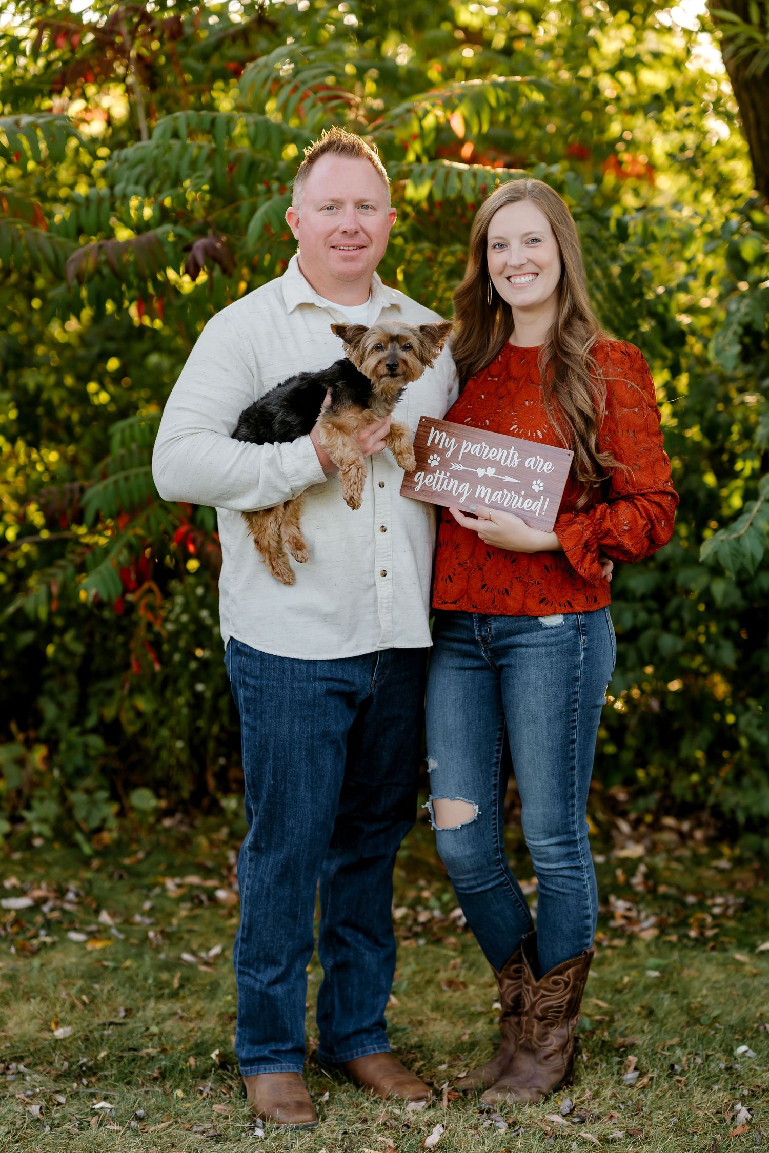 Couple in casual fall outfits with a small dog poses together in front of autumn foliage during outdoor photoshoot.