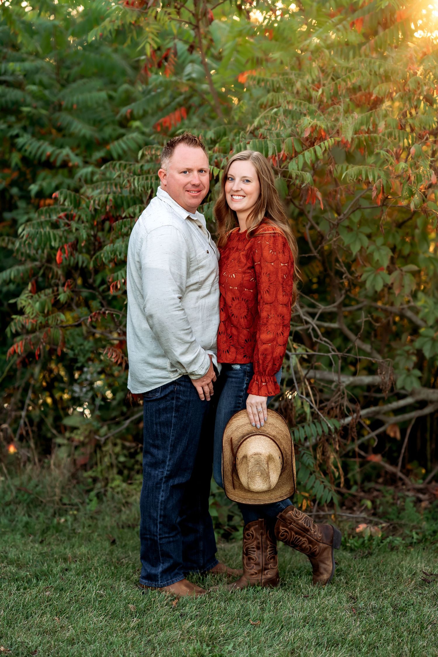 A couple in casual western attire shares a romantic moment against a backdrop of autumn foliage at sunset.