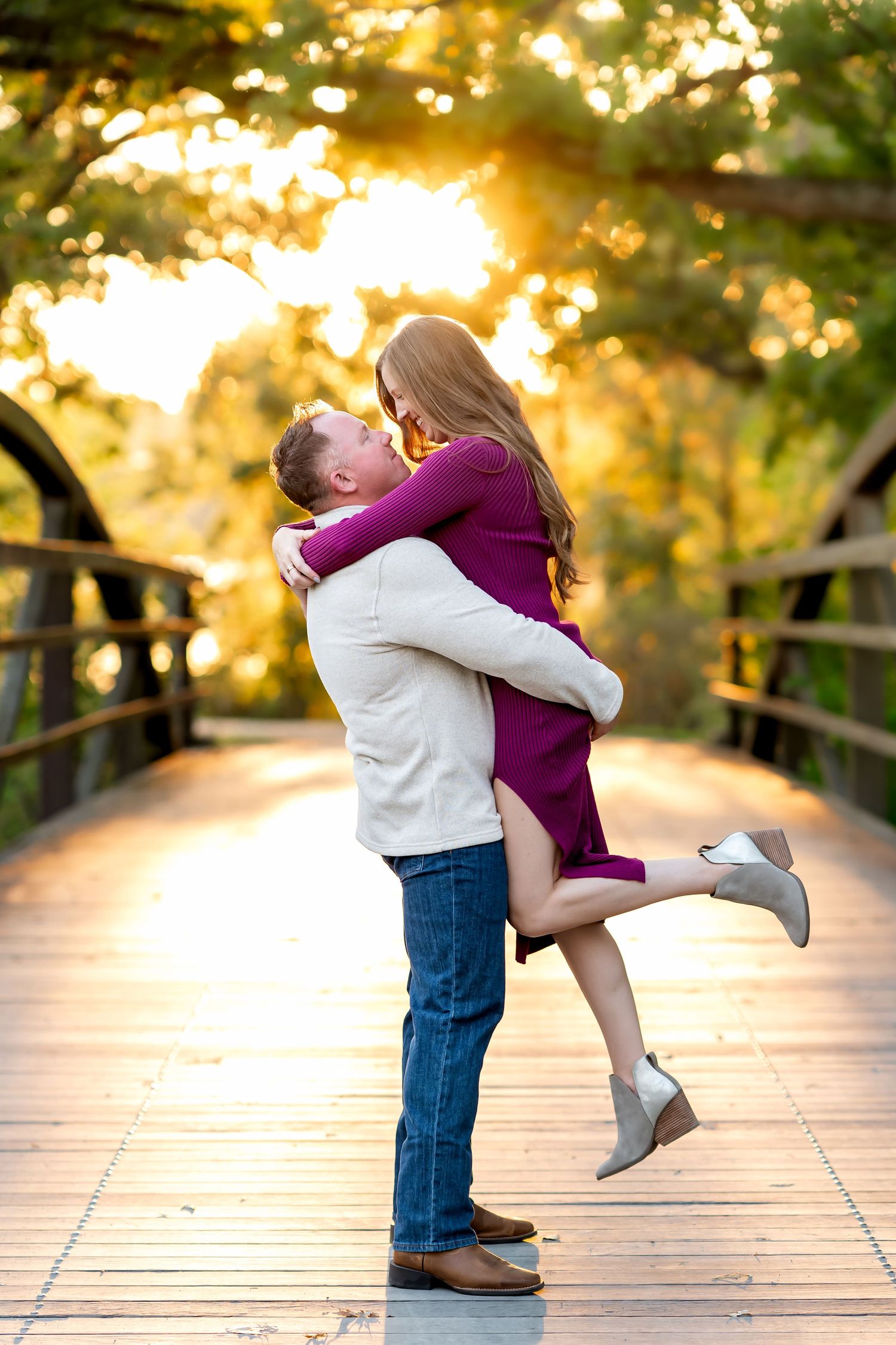 A couple shares a romantic embrace on a wooden bridge during golden hour sunset with autumn trees in background.