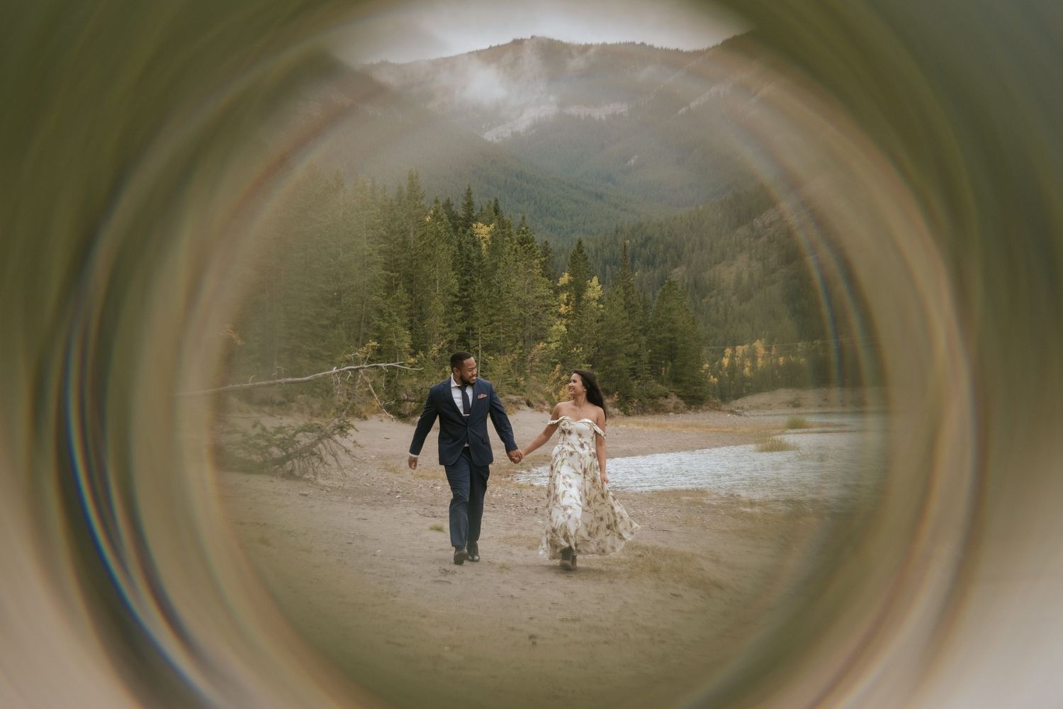 Couple walking hand in hand along a scenic mountain path surrounded by evergreen trees and misty peaks.