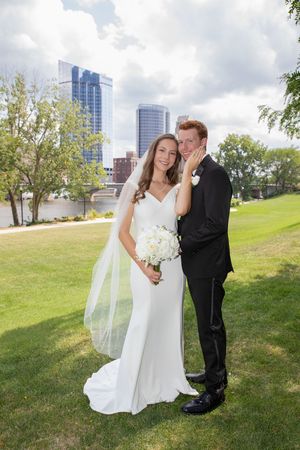A couple in wedding attire share a tender moment on a grassy hill with a city skyline visible in the background.